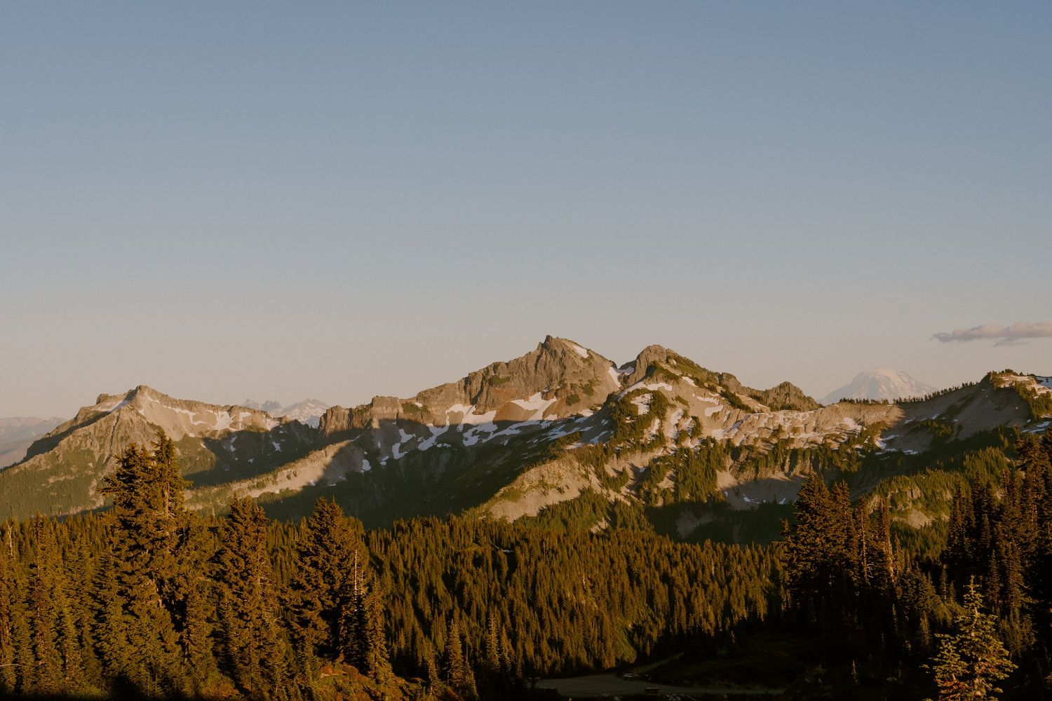 Golden hour light illuminates dense evergreen forests below rugged snow-capped mountain peaks under a clear blue sky.