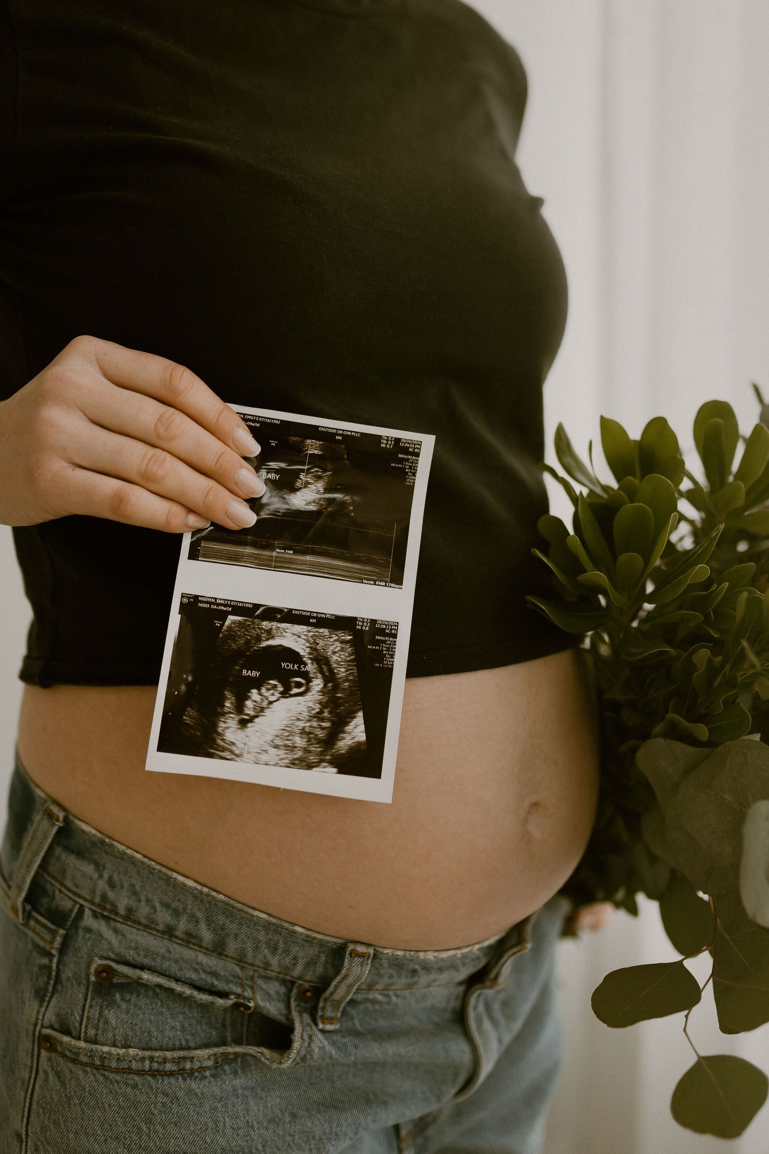 A series of ultrasound photos held against a pregnant belly wearing a black top and olive jeans.