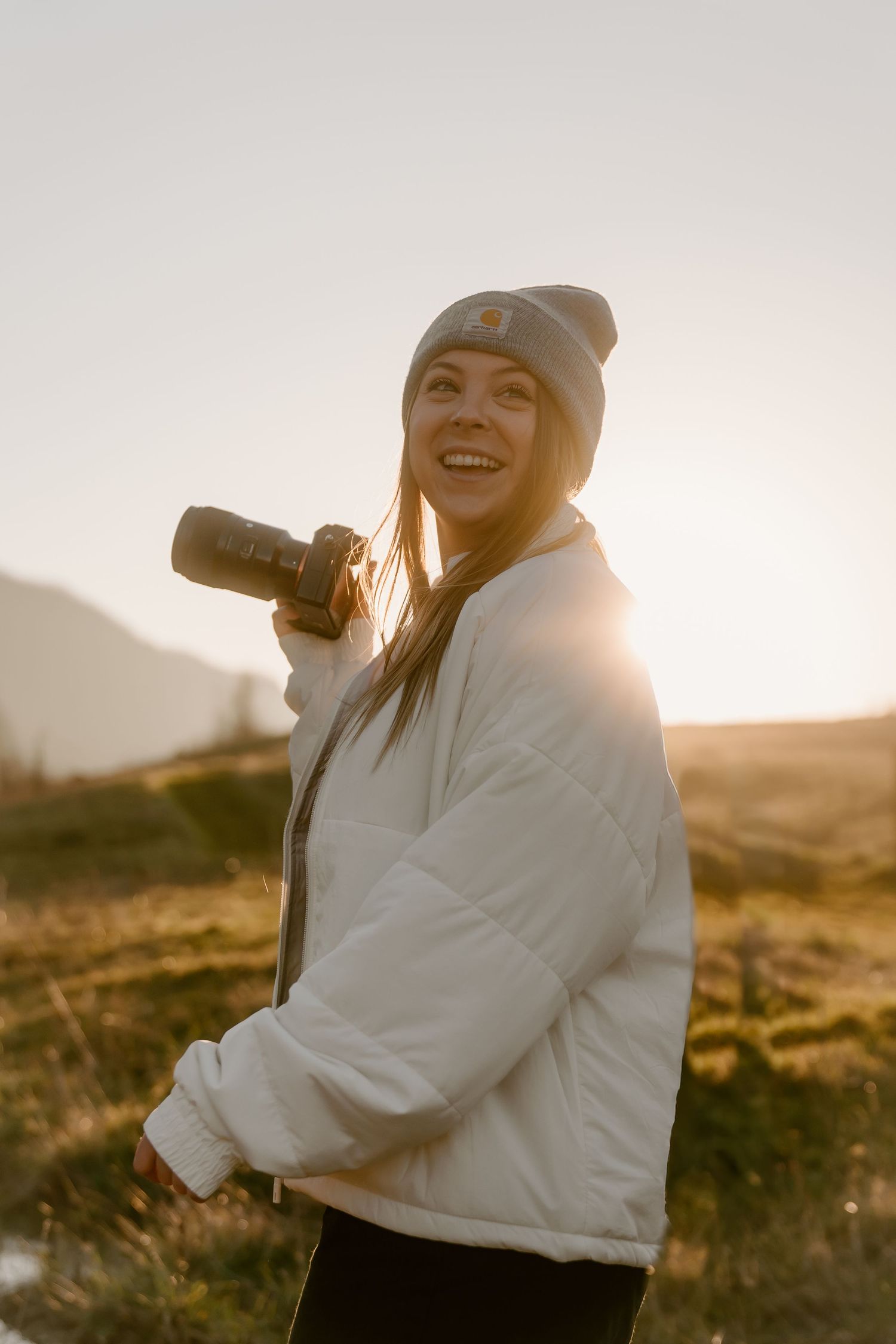 Smiling woman holding a camera outdoors at golden hour, wearing a white puffer jacket and gray beanie hat.