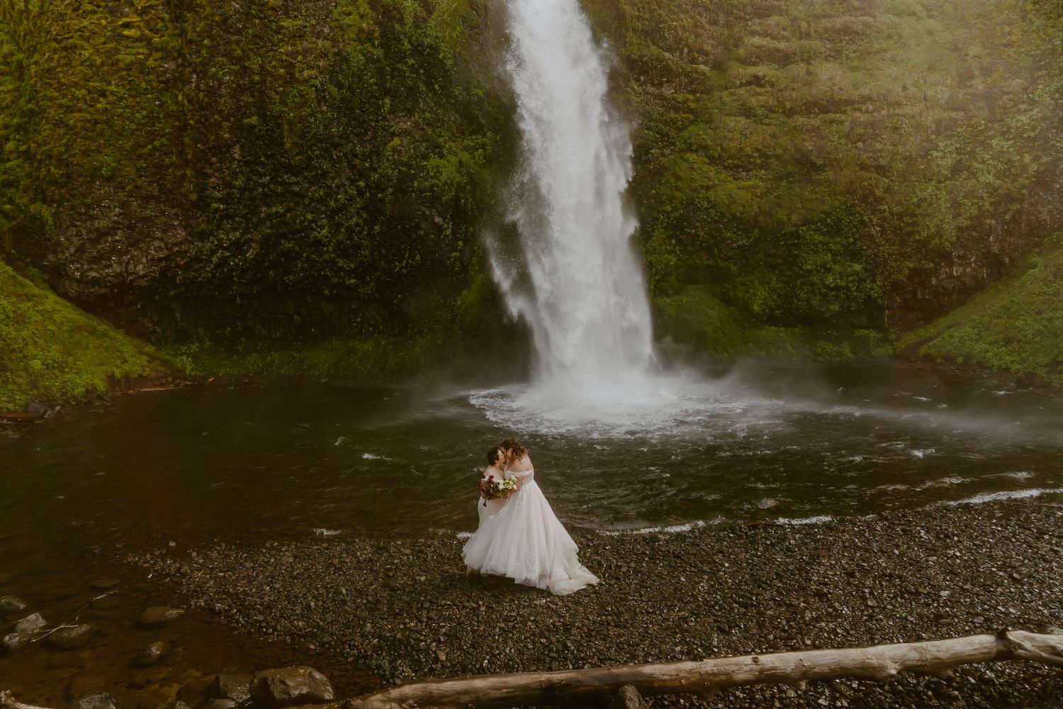Bride and groom embrace before a stunning waterfall surrounded by lush greenery in an outdoor wedding photo.