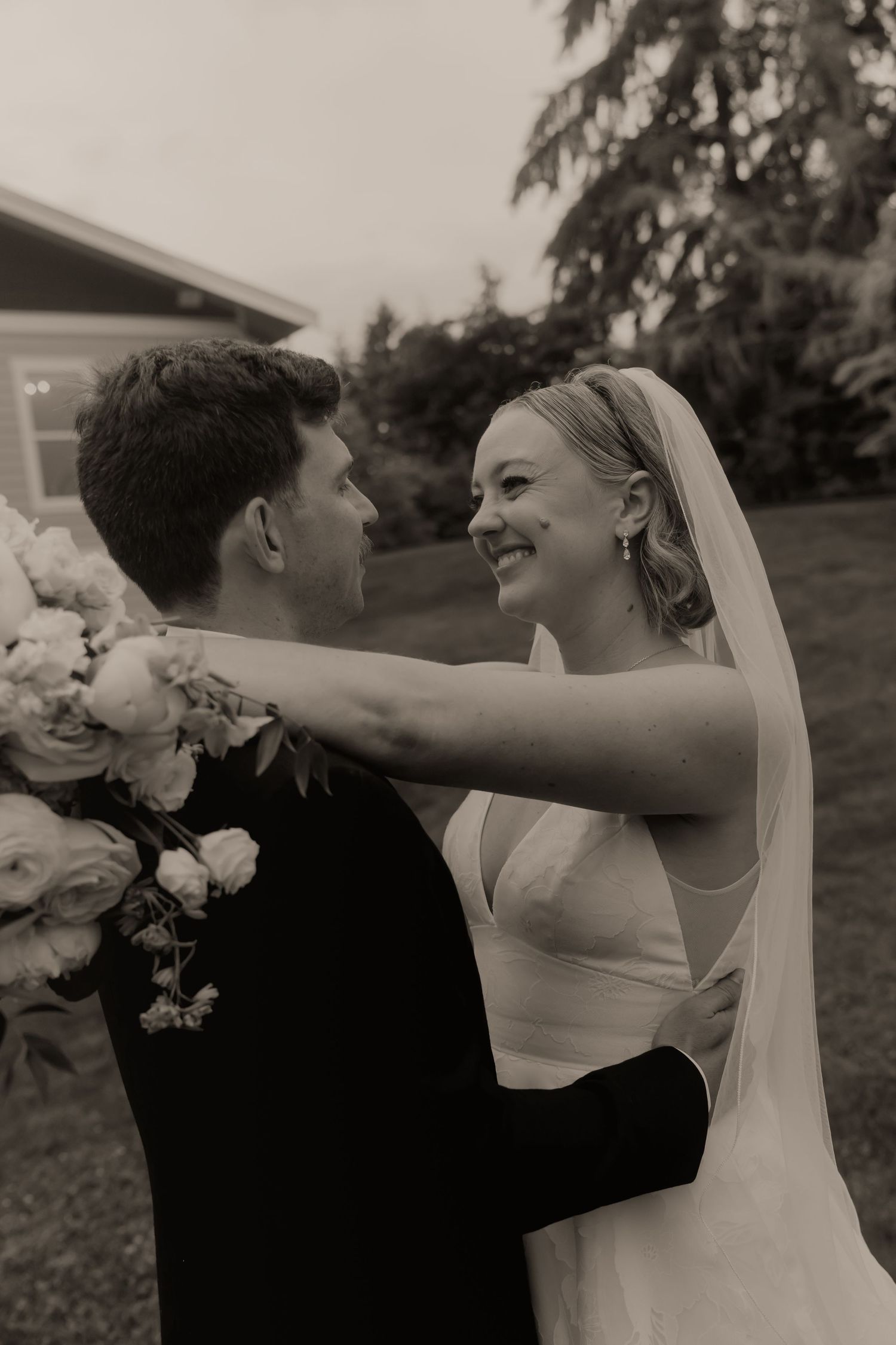 Black and white wedding photo of smiling bride in veil embracing groom outdoors with floral bouquet.
