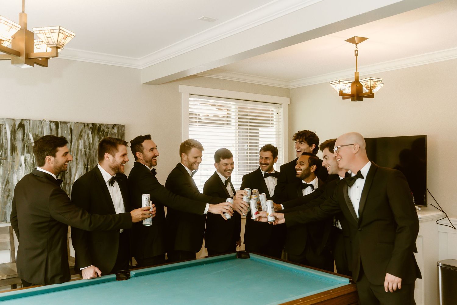 Groomsmen in black tuxedos toasting with drinks around a pool table in an elegant room.