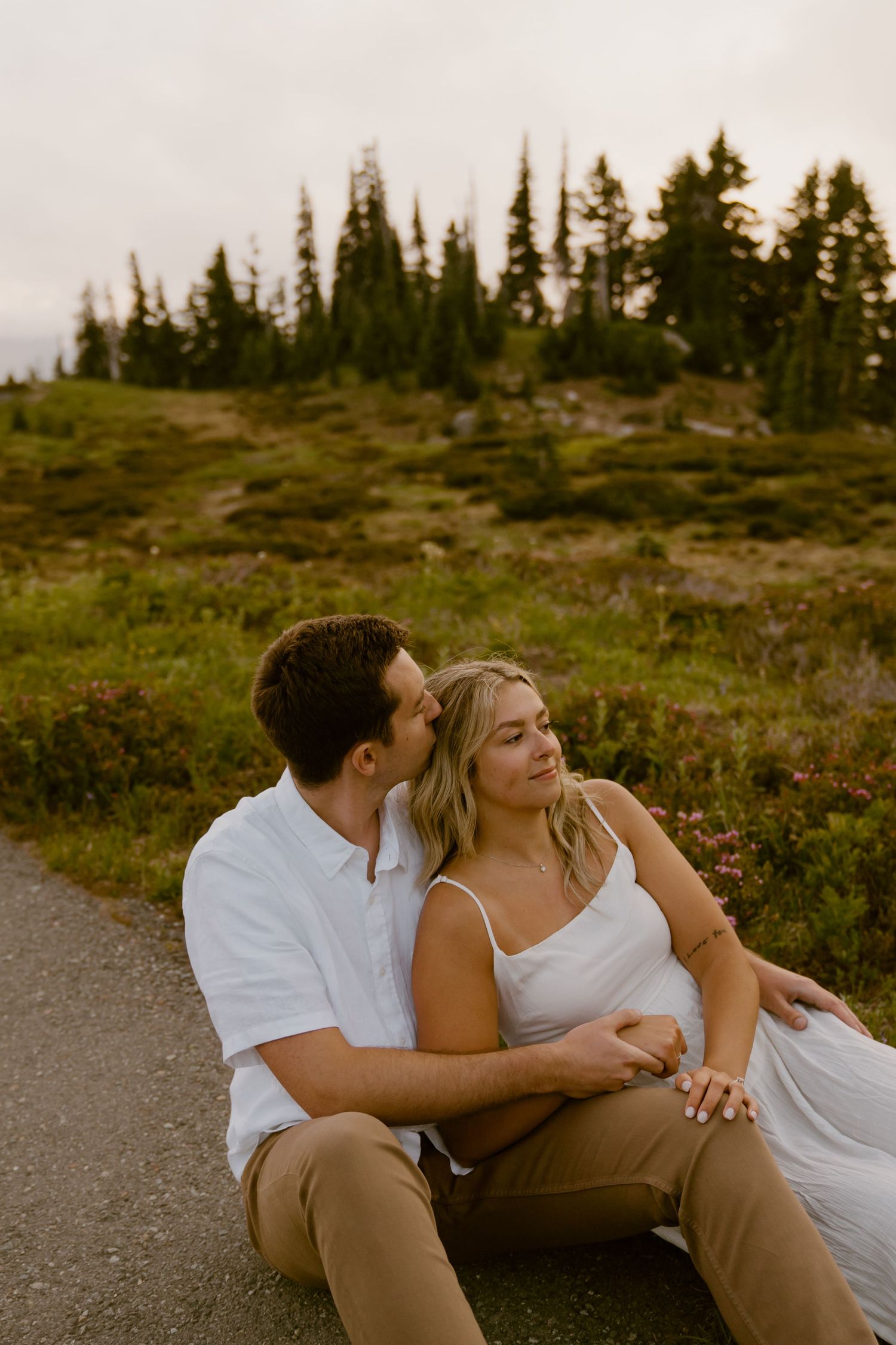 Couple relaxing on path amid evergreen trees during mountain sunset photoshoot.