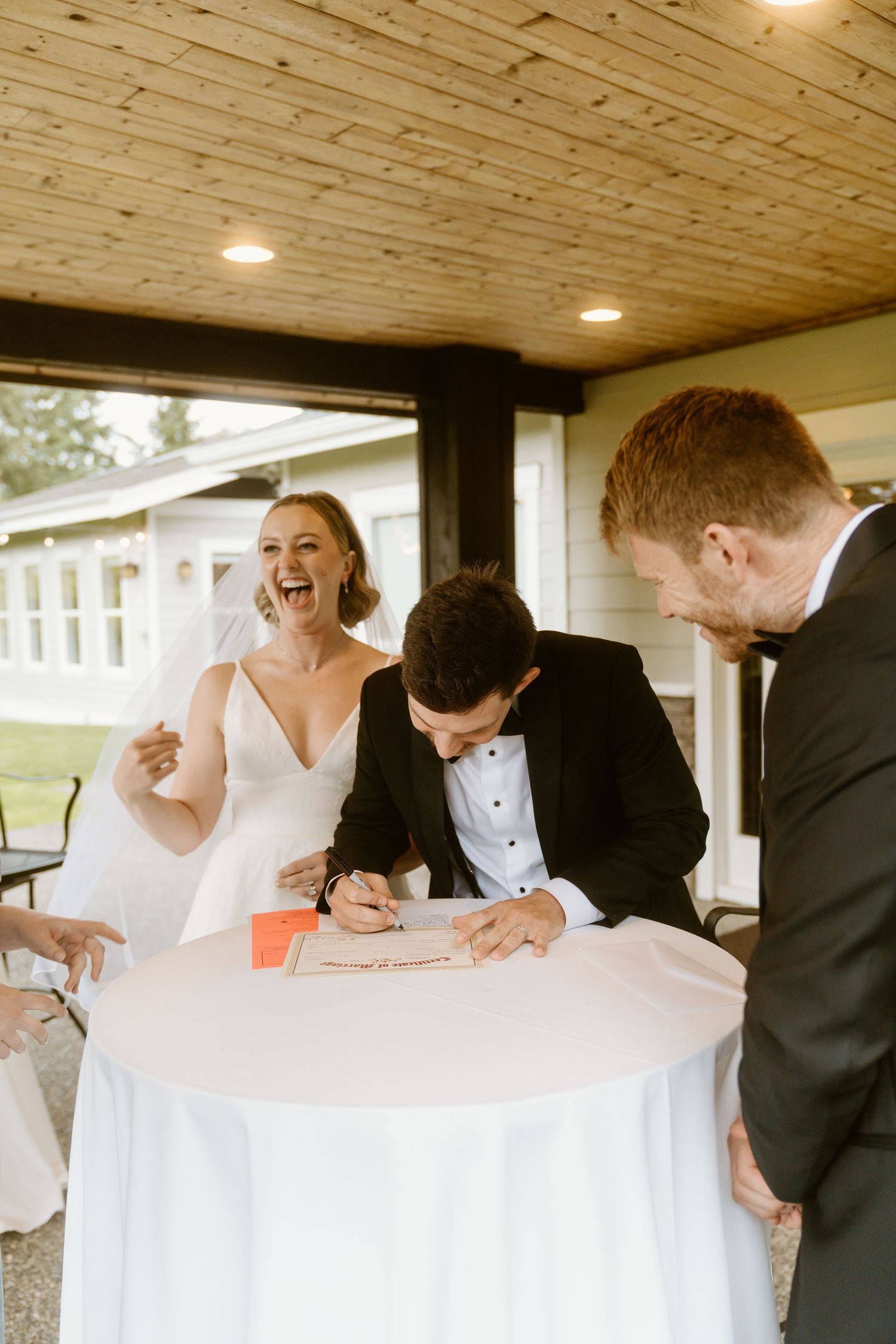 Bride laughing joyfully as groom signs wedding documents at a white table, officiant standing nearby at outdoor venue.