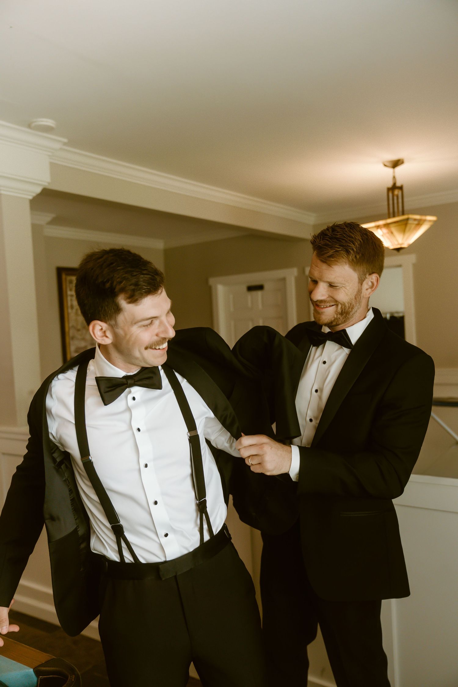 Two groomsmen in black tuxedos with bow ties laugh while helping each other get dressed.