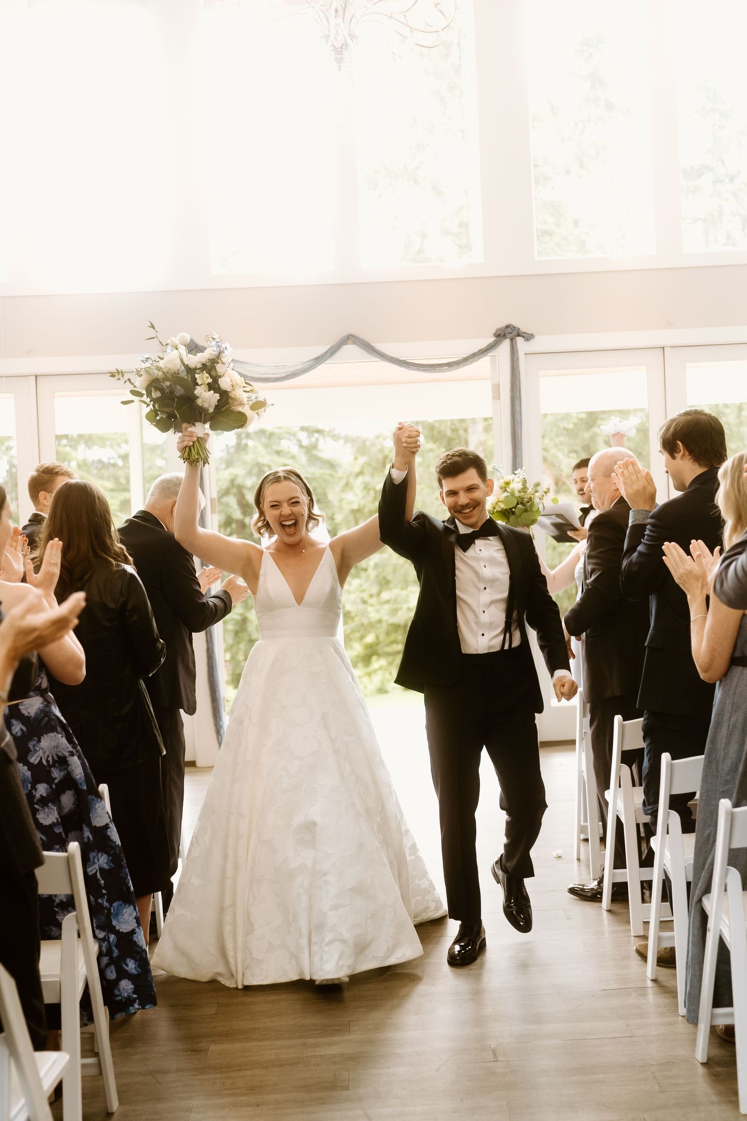 Joyful bride and groom raising hands while walking down the aisle after their indoor wedding ceremony.
