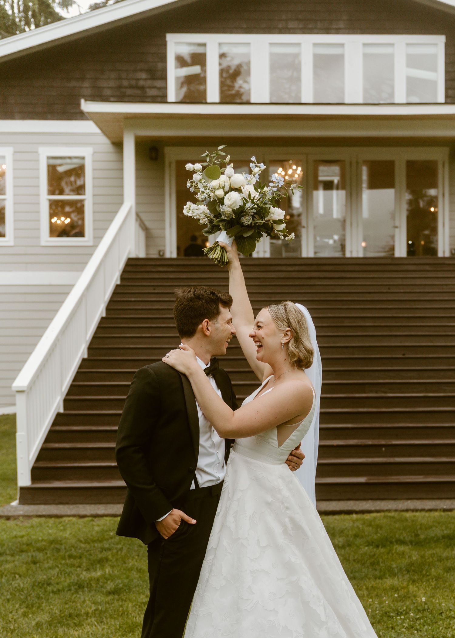 Joyful bride raises bouquet of white flowers while laughing with groom outside wedding venue.