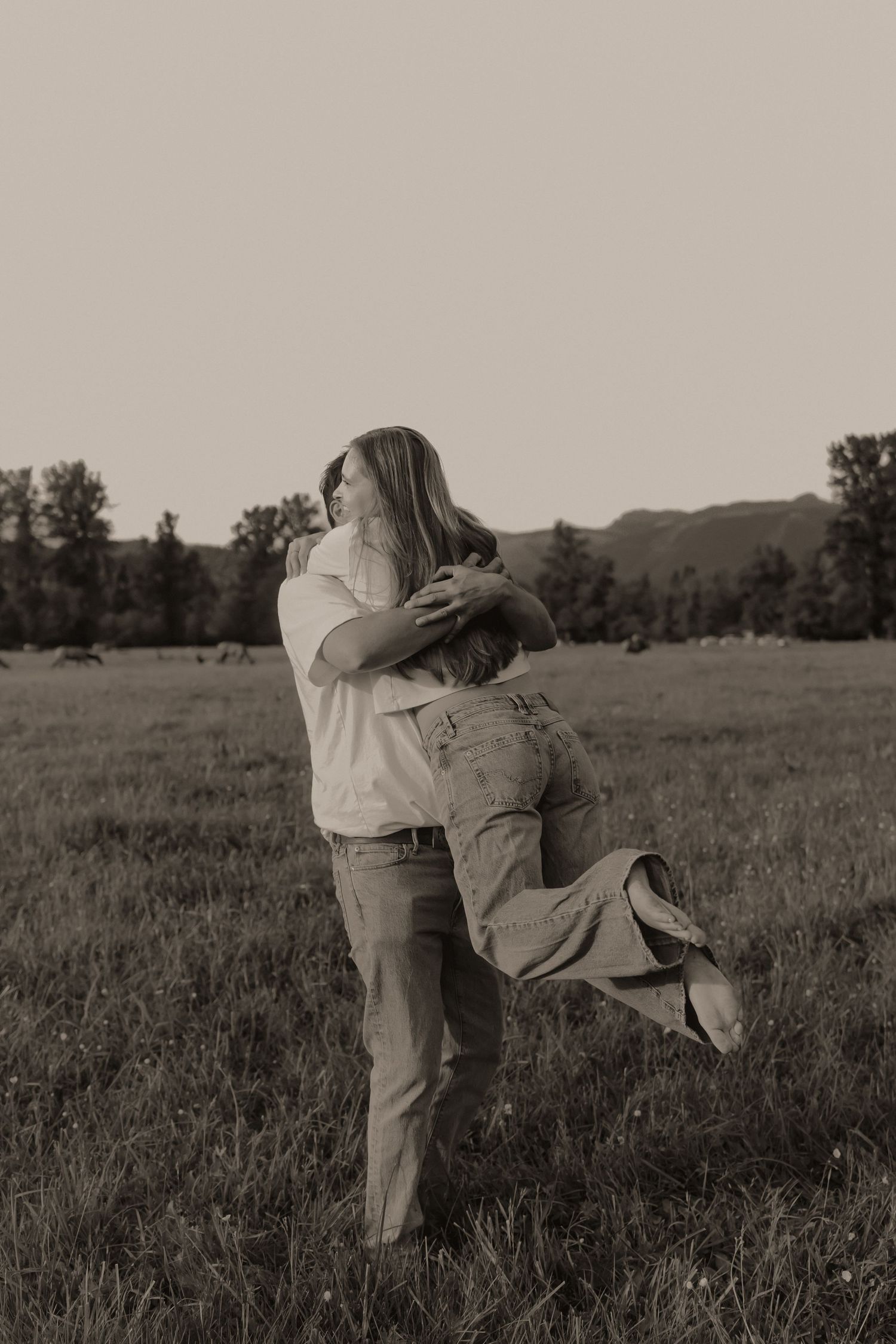 Couple embracing romantically in a grassy field with mountains in the background, sepia-toned photo.