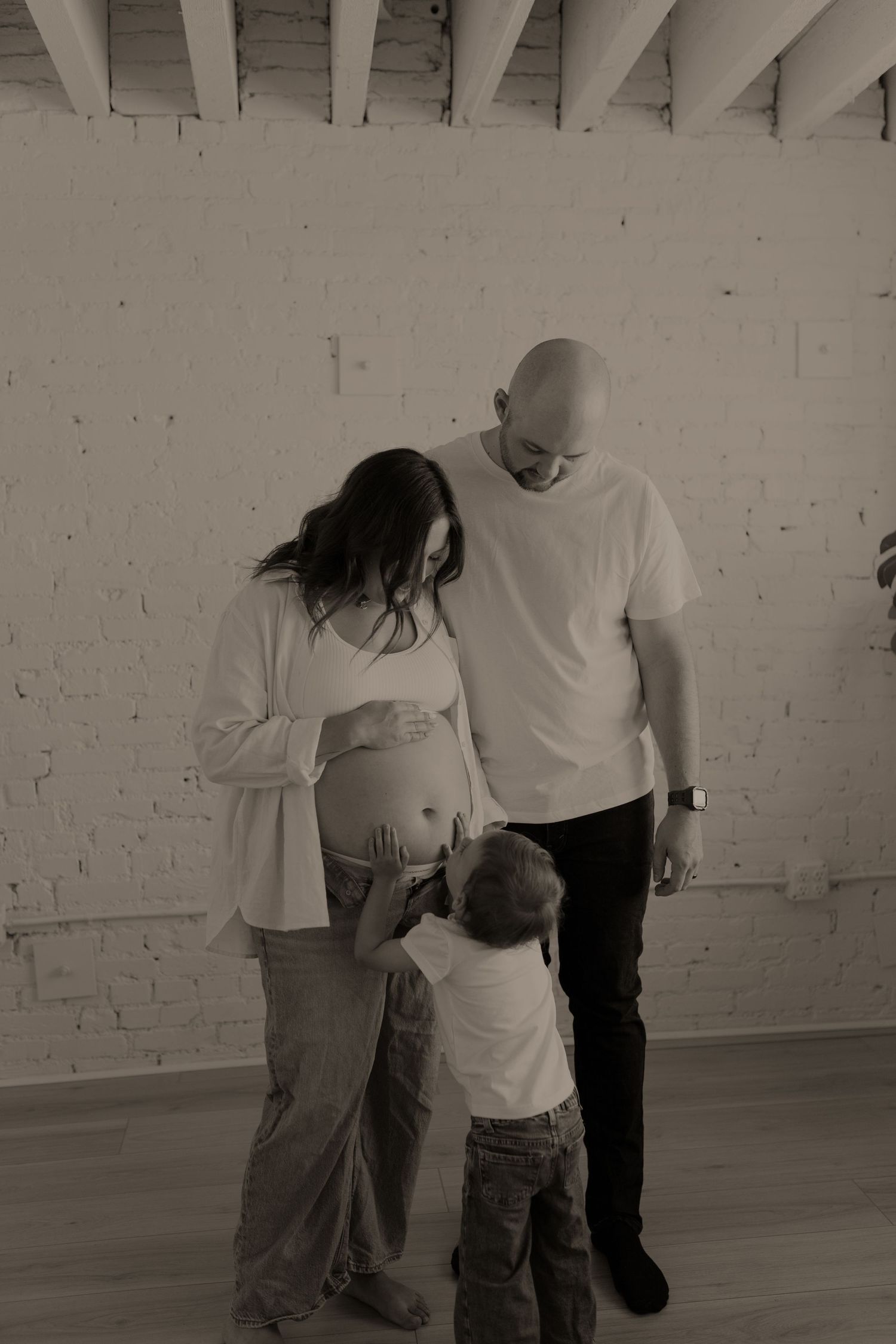 Black and white maternity photo of pregnant woman, bald man, and toddler child touching baby bump in bright studio.