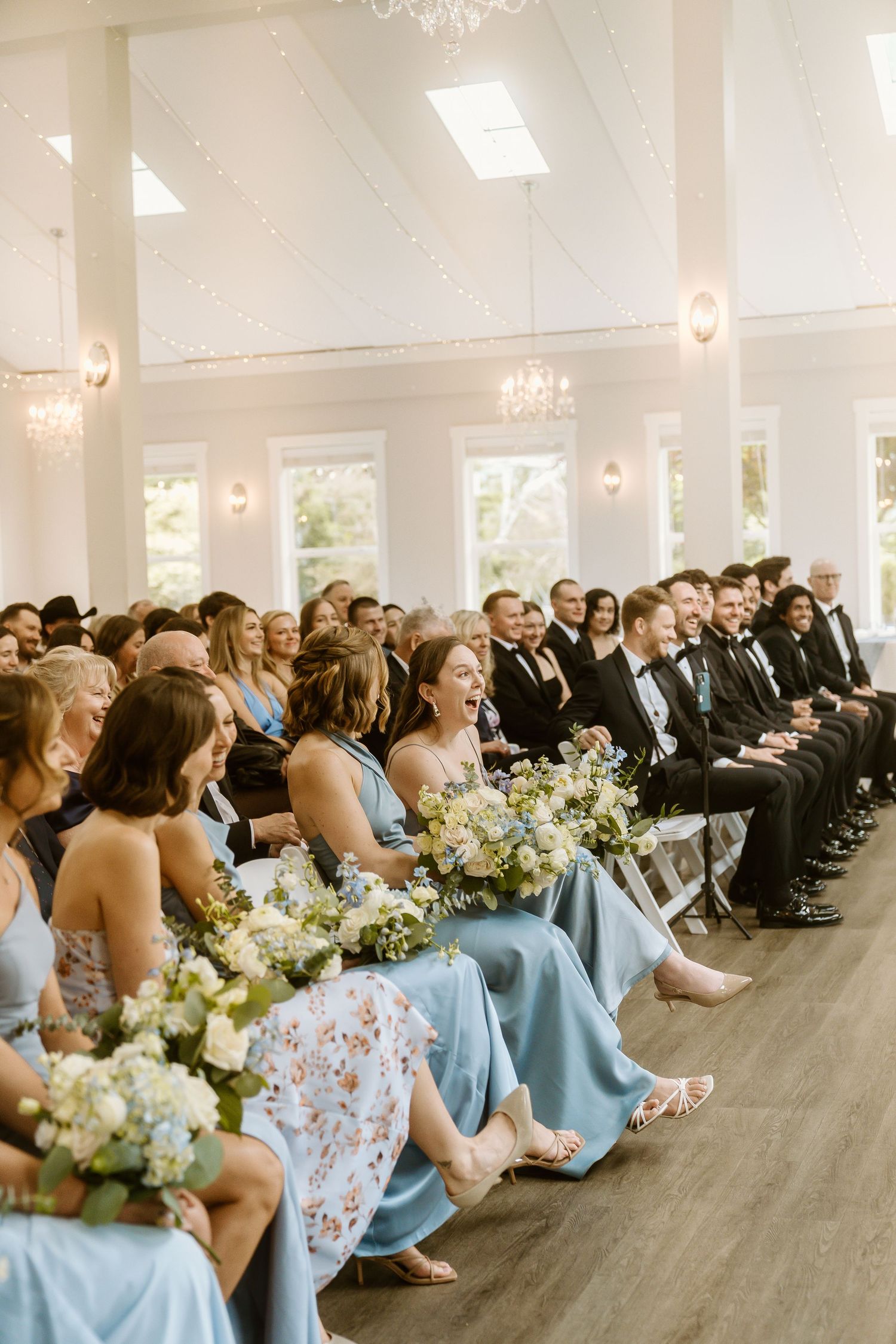 Bridesmaids in blue dresses holding white floral bouquets seated at an elegant indoor wedding ceremony.