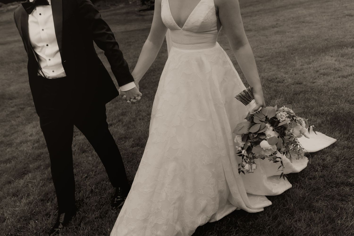 Black and white photo of a bride in a white gown holding a bouquet and groom in a tuxedo holding hands outdoors.