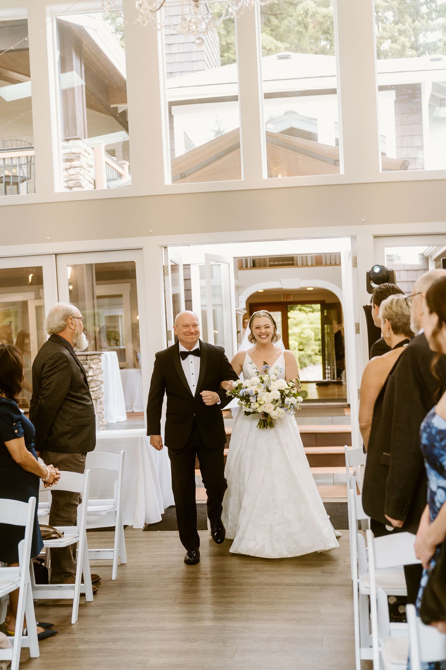 Bride walks down the aisle escorted by her father in elegant indoor wedding venue with tall windows.