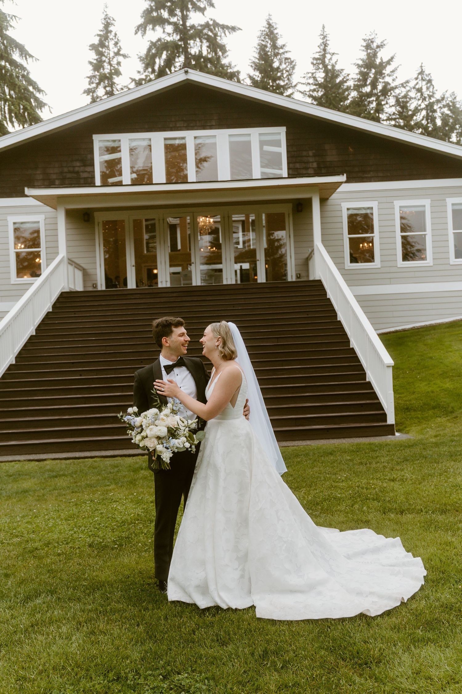 Bride and groom smiling at each other on green lawn before elegant venue with grand staircase and tall evergreen trees.