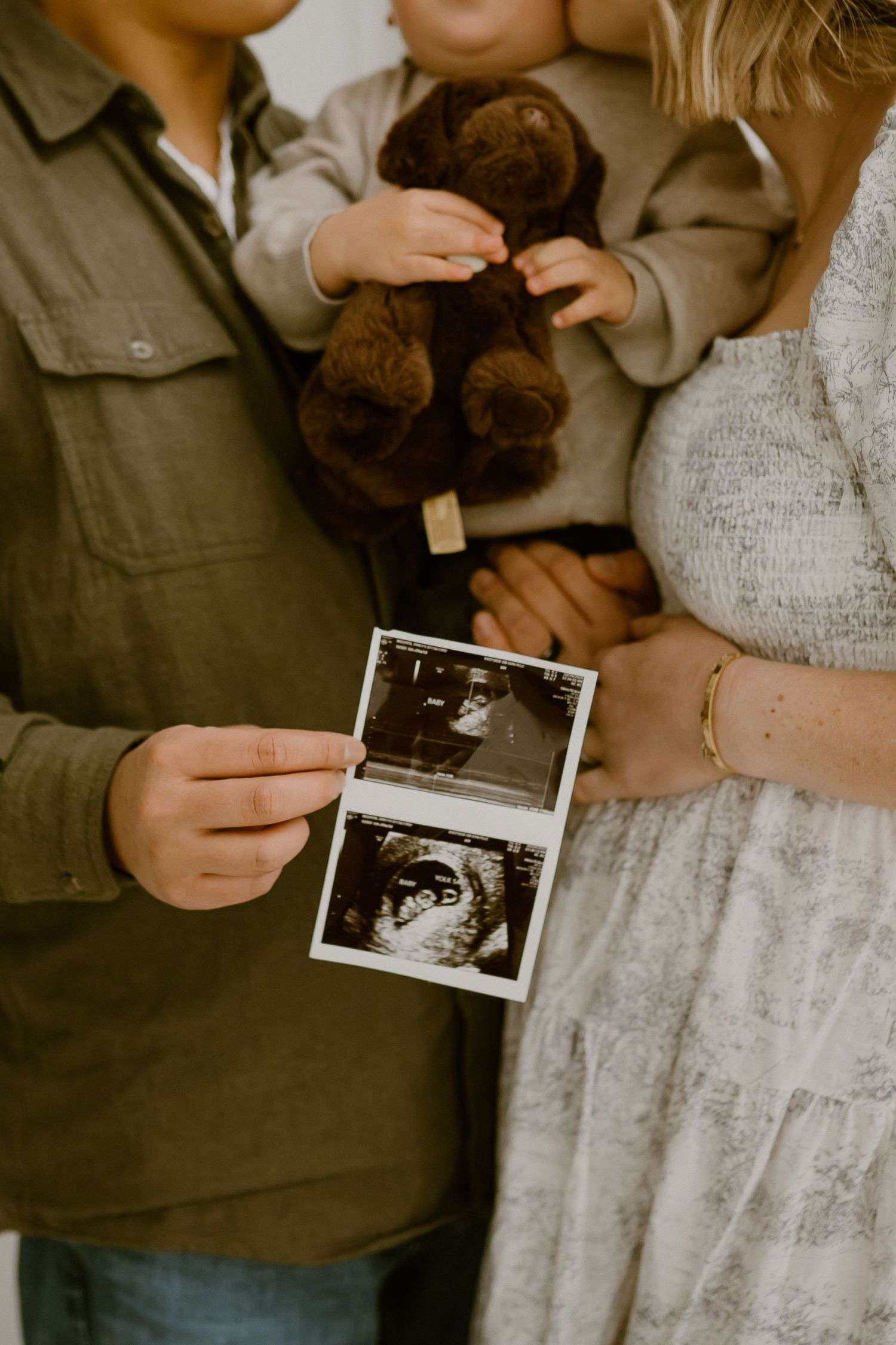 A couple holds ultrasound photos and a teddy bear while announcing their pregnancy with a toddler nearby.