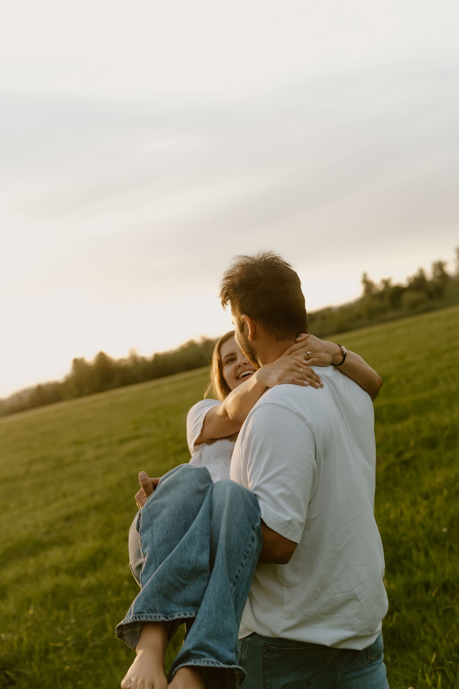 Man carrying woman in his arms in a green field at sunset, both wearing white shirts and jeans.