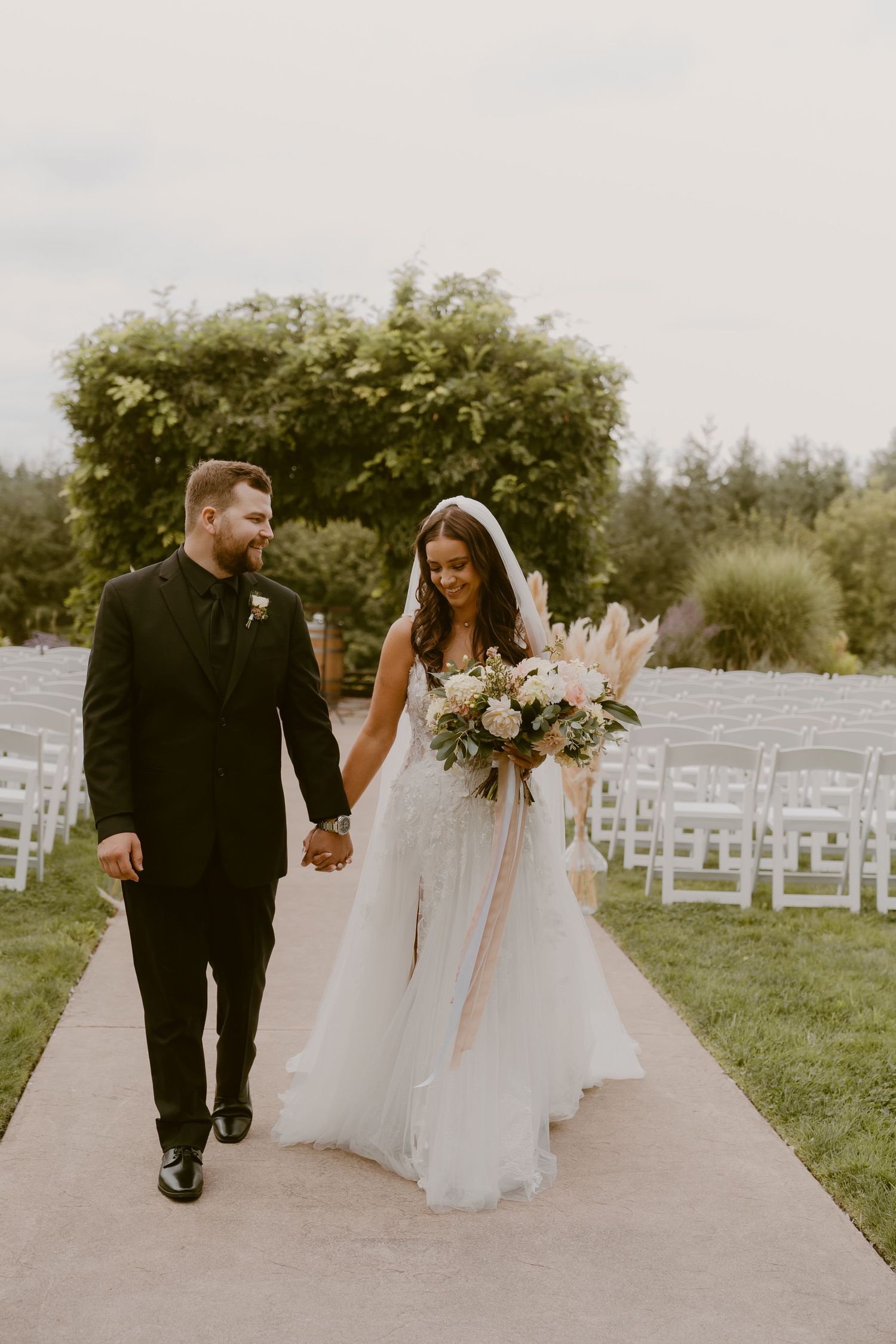 Newlyweds walk hand in hand on outdoor path with white chairs and greenery in background.