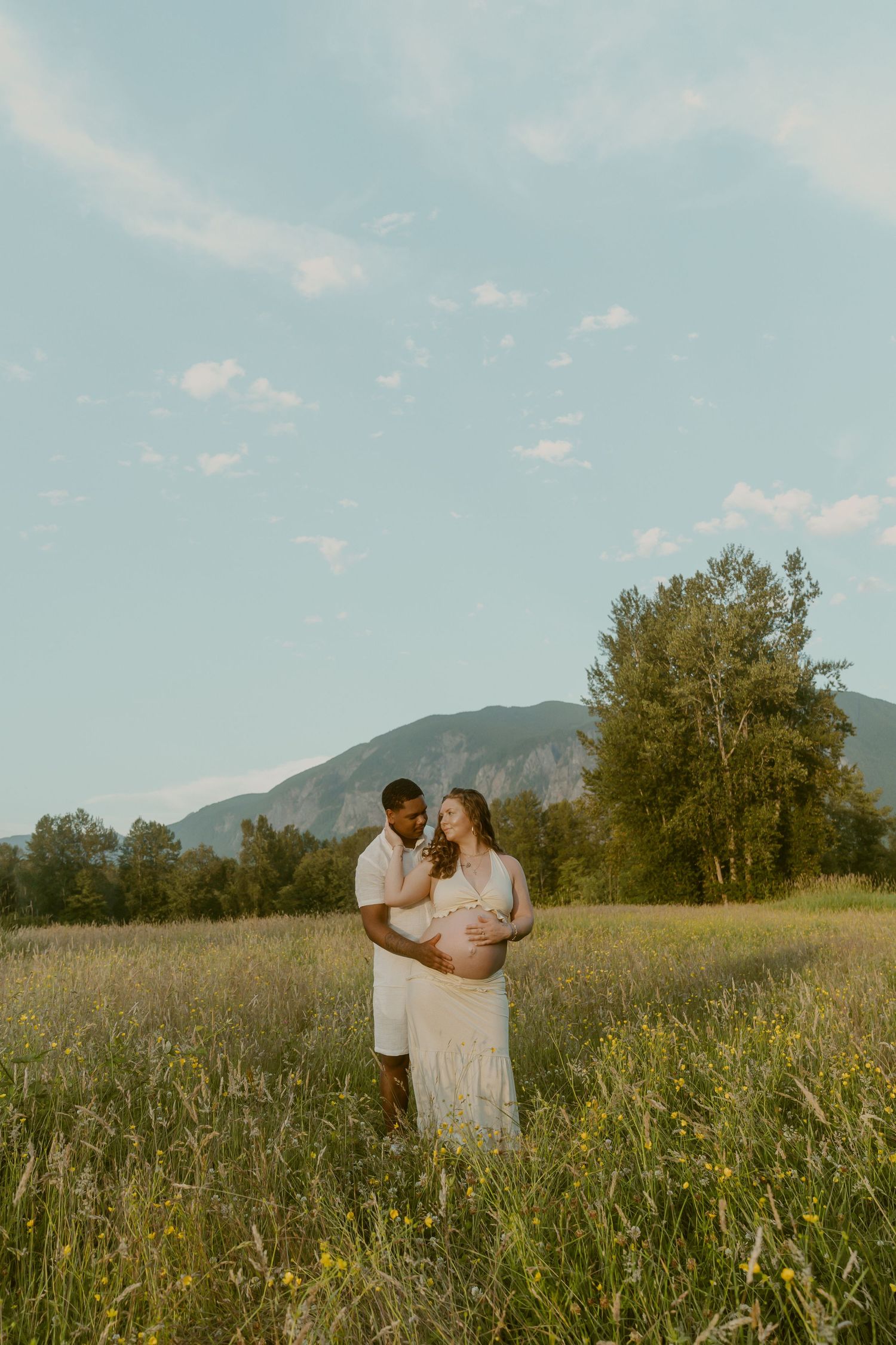 Couple in white standing in a sunlit meadow with mountain backdrop during a maternity photoshoot.