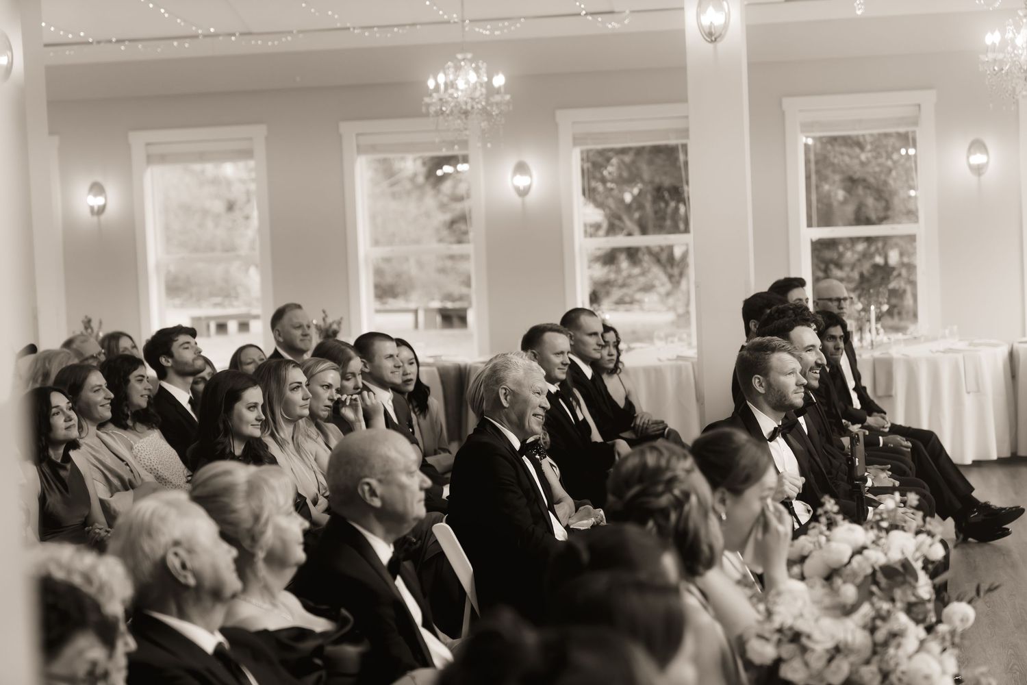 Elegantly dressed wedding guests seated in a grand ballroom during a wedding ceremony, captured in black and white.