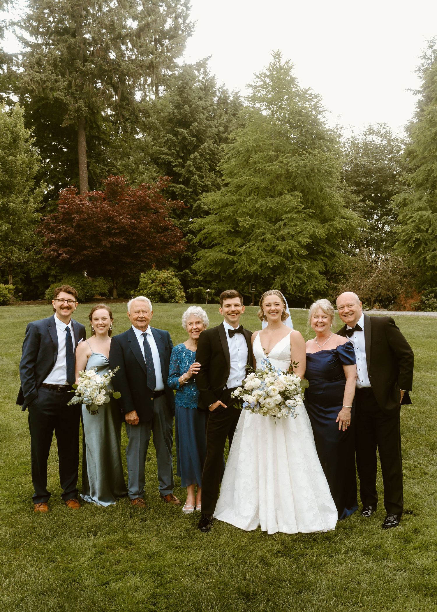Wedding party poses outdoors on green lawn with tall trees, bride in white gown holding bouquet with groom in tuxedo.