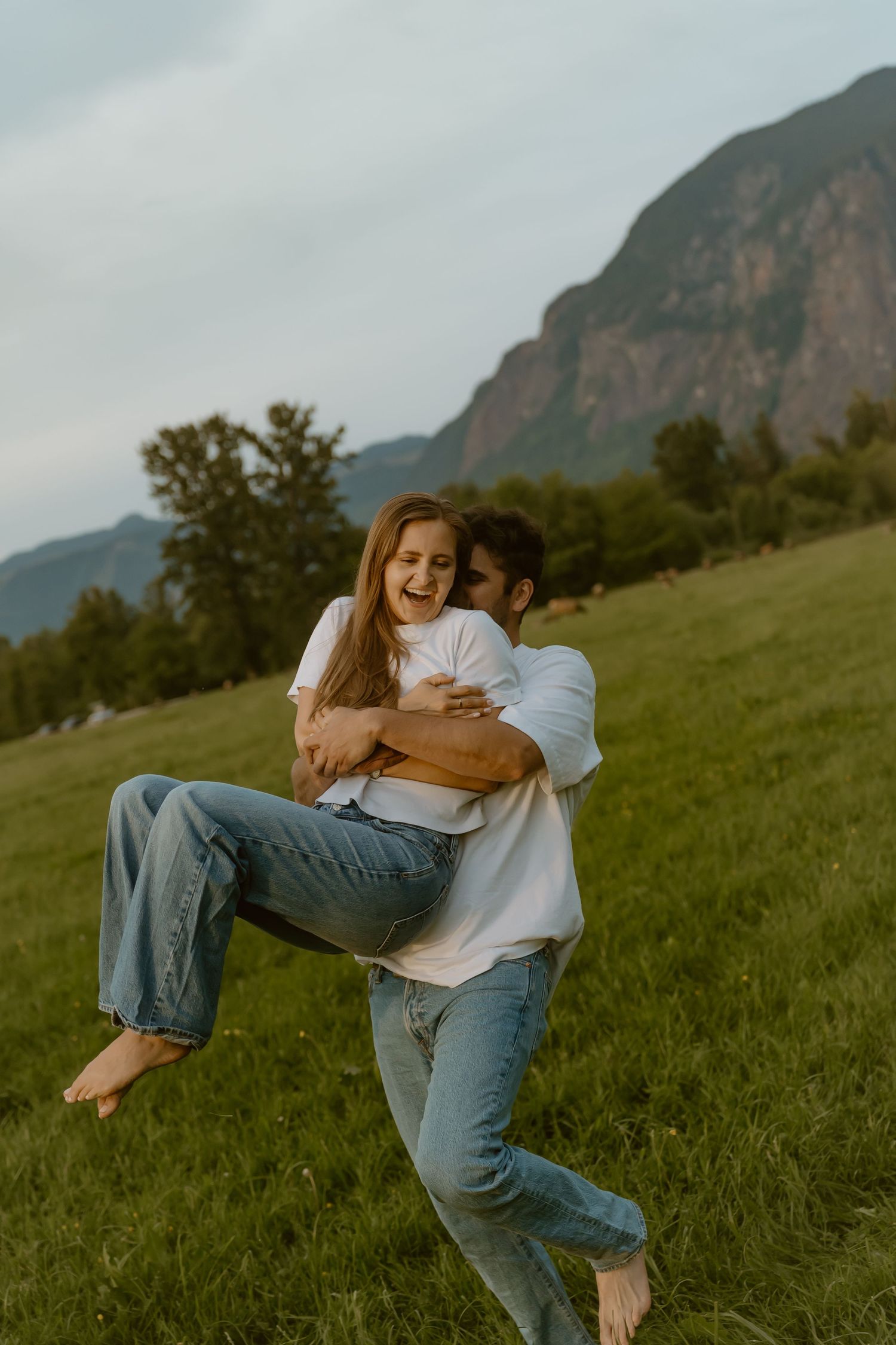 Happy couple in white shirts and jeans playing in a green meadow with mountains in the background.