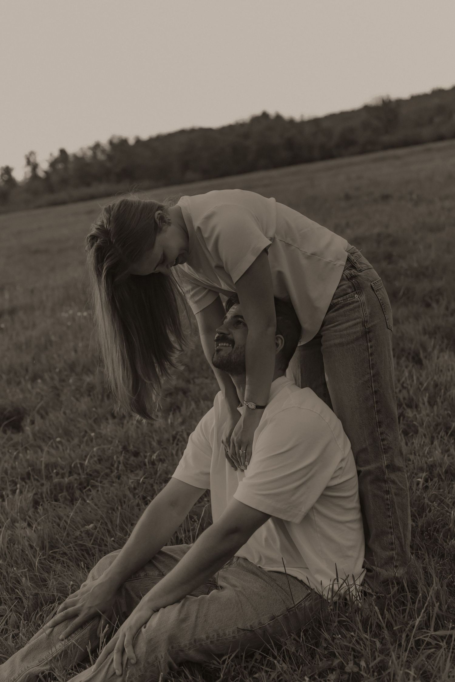 A sepia-toned photo of a couple in a field, with a woman leaning over a man sitting on the grass.