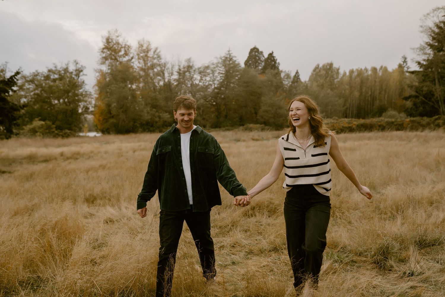 A joyful couple holds hands and laughs while walking through a golden autumn meadow surrounded by tall trees.