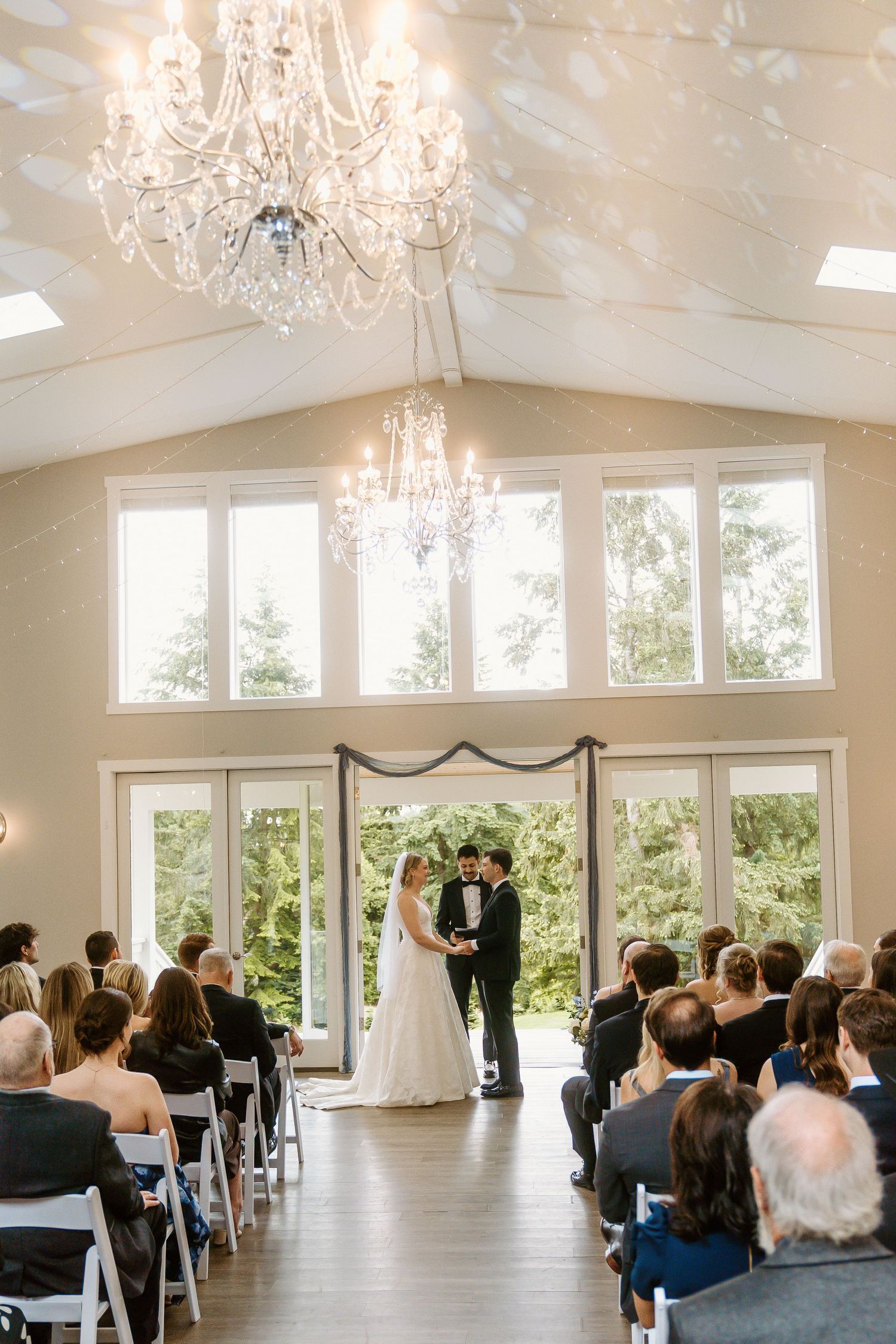 Elegant indoor wedding ceremony with crystal chandelier, bride and groom at altar, guests seated in white chairs, large windows backdrop.