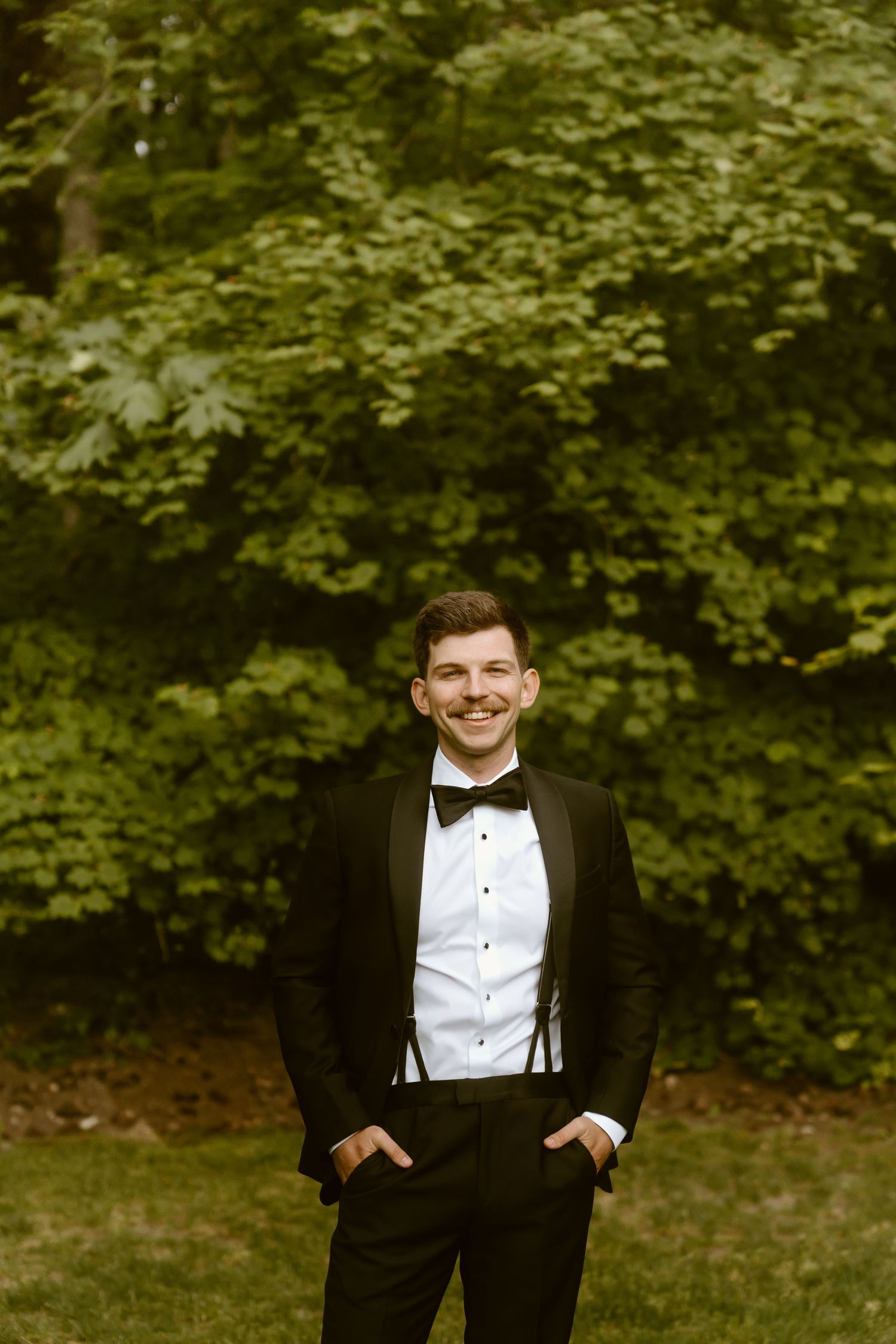 Smiling groom in black tuxedo with bow tie and suspenders standing outdoors against lush green trees.