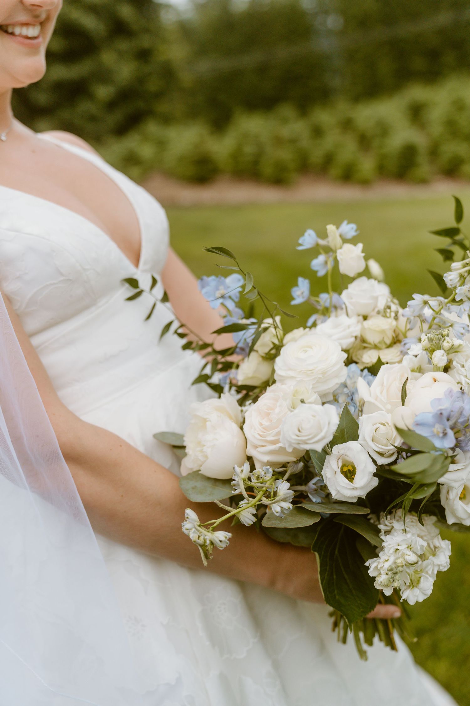 Close-up of bride holding lush bouquet of white roses and blue flowers in garden setting.
