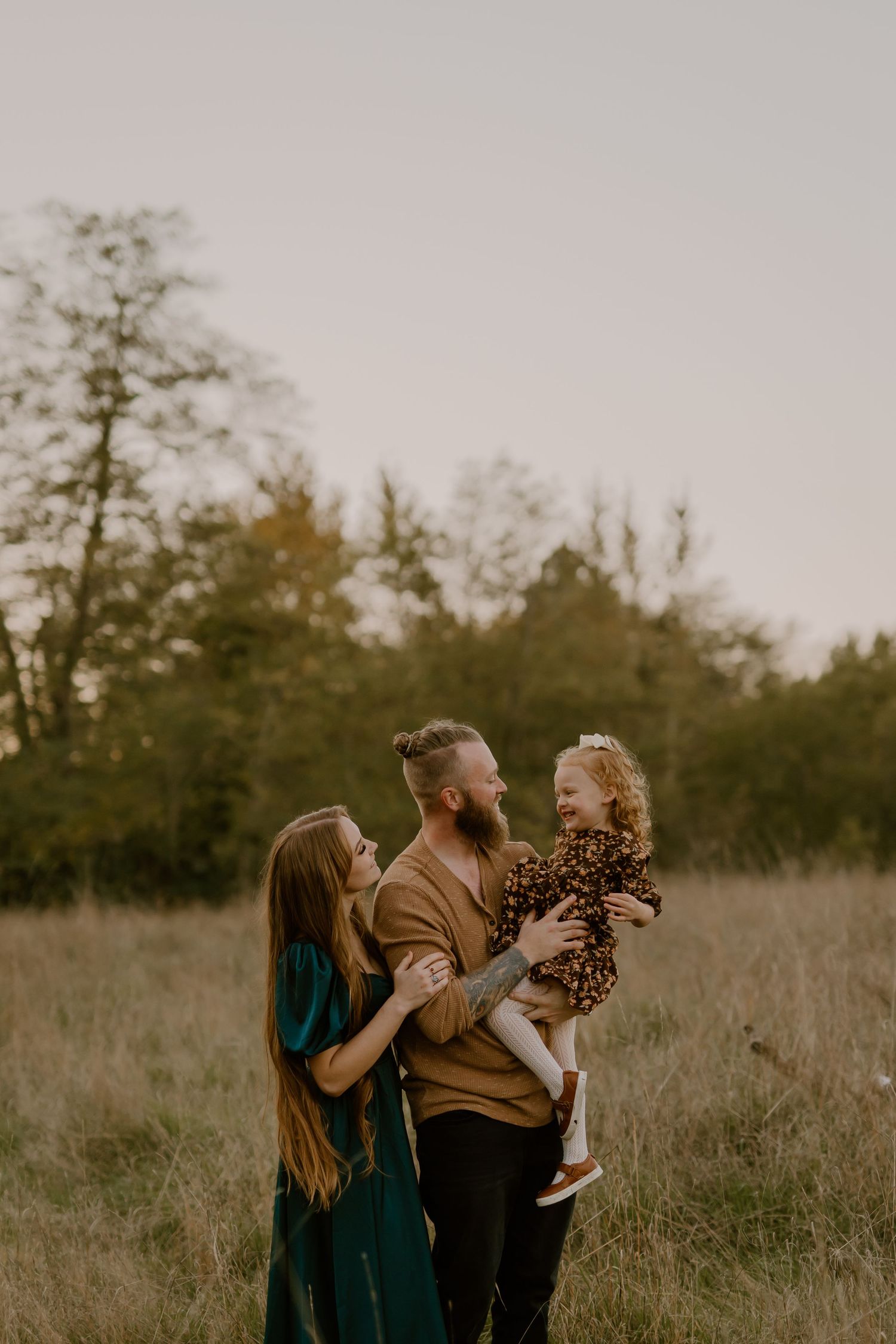 A happy family of three poses in a golden field, with parents smiling at their laughing toddler daughter.