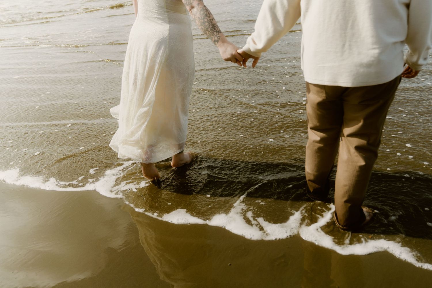 Barefoot couple in wedding attire holding hands while walking along a sunlit beach shoreline.