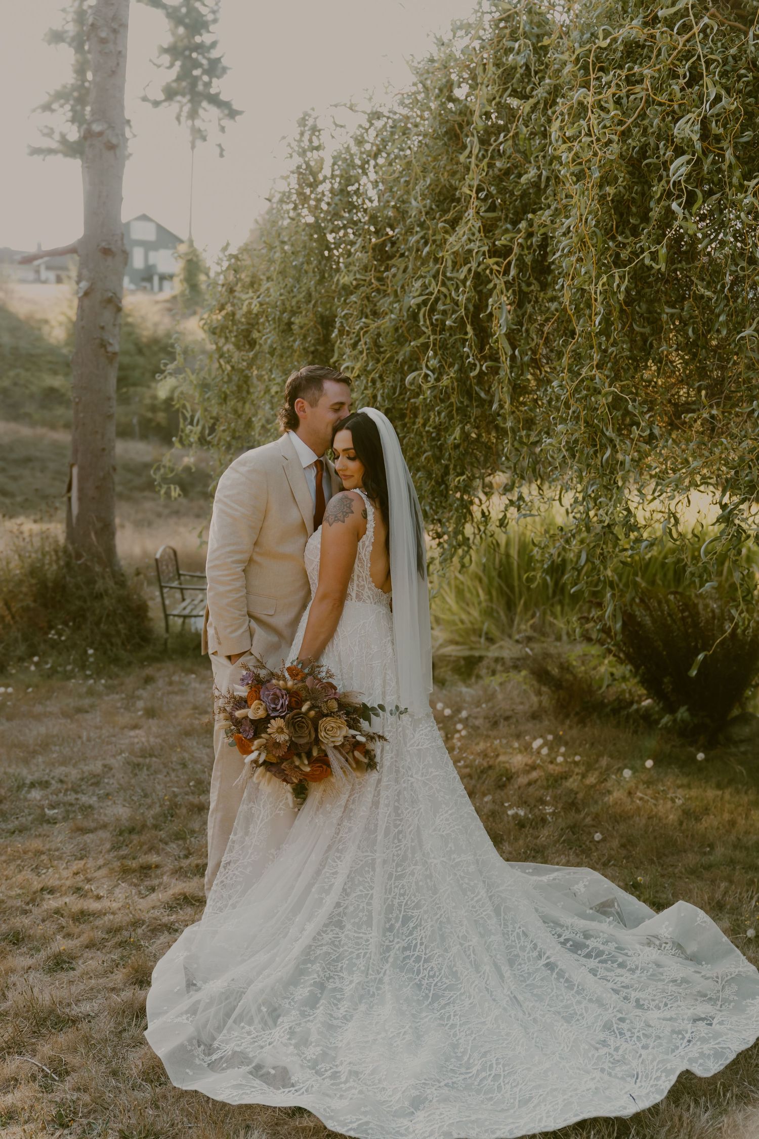 Newlywed couple embraces in a romantic outdoor setting with flowing white lace wedding dress and autumn-toned bouquet.