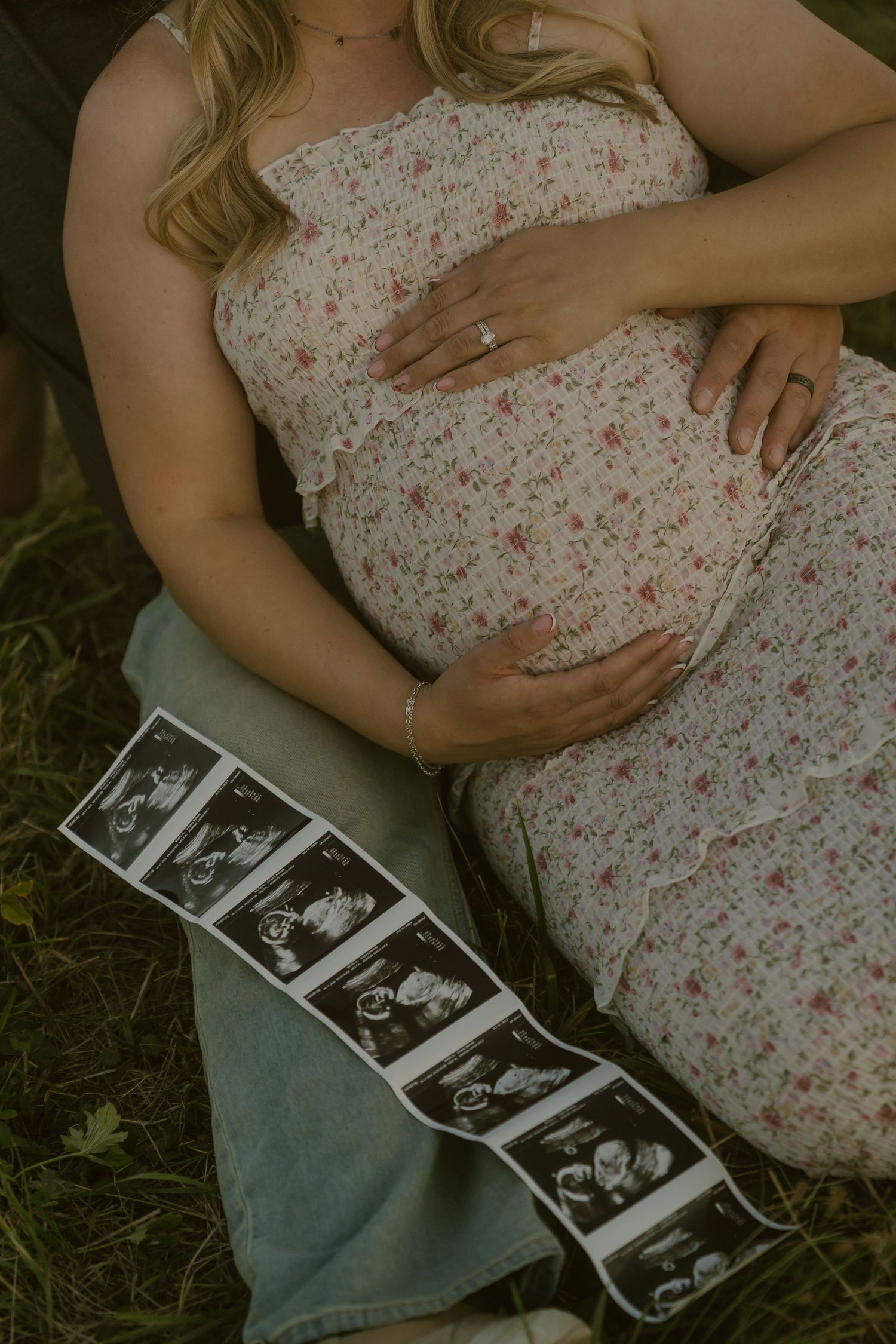 Pregnant woman in a floral dress cradling her baby bump while sitting on grass with a strip of ultrasound photos draped across her lap.