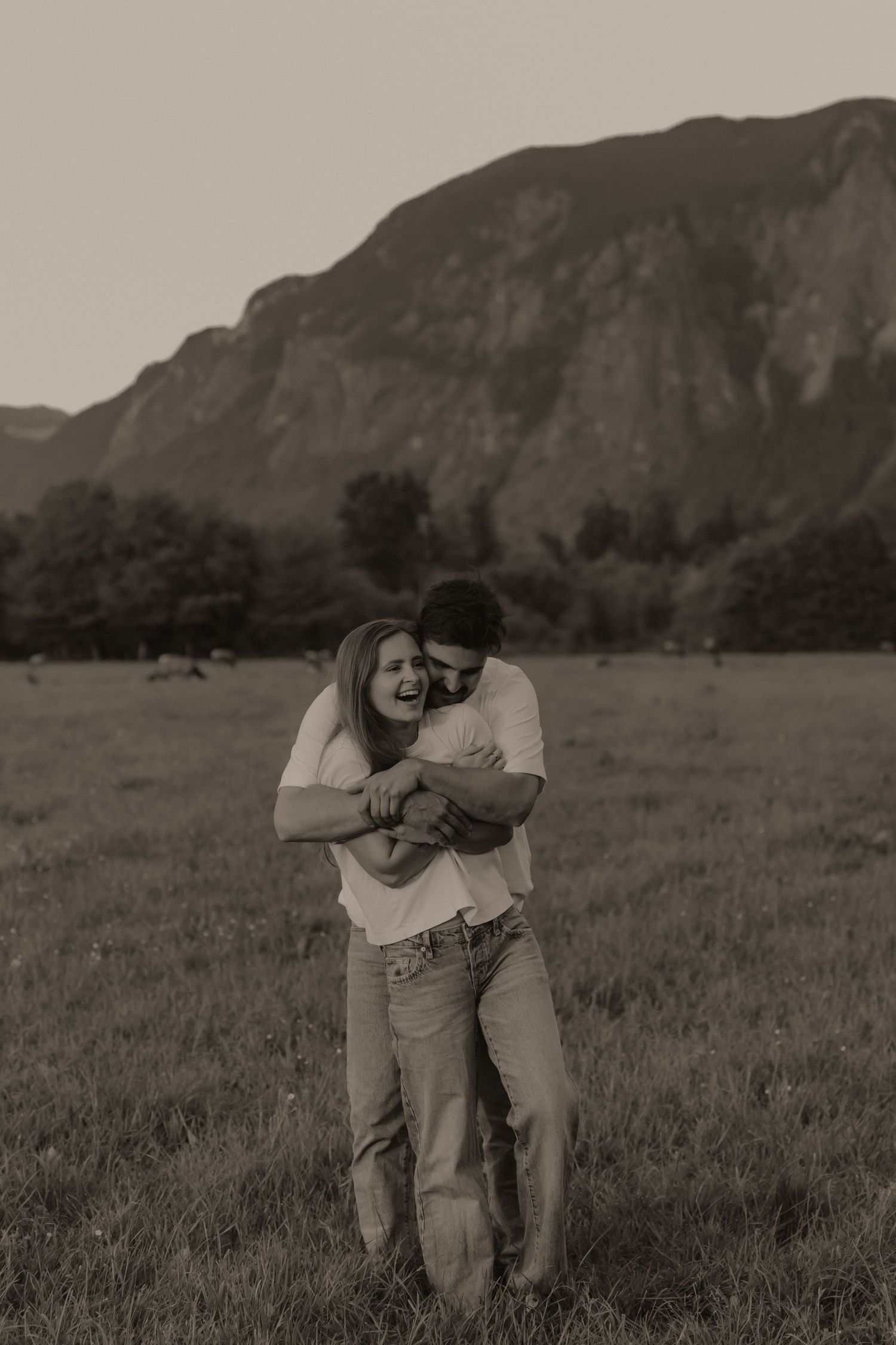 Couple laughing and embracing in a grassy field with a mountain backdrop in a sepia-toned outdoor photo.