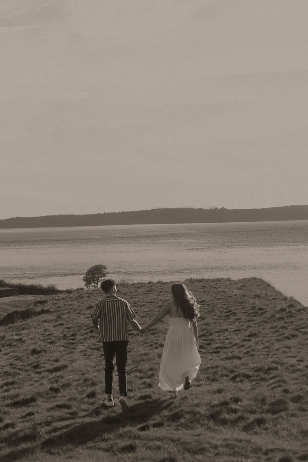 Couple holding hands walking toward a serene lake in a sepia-toned romantic outdoor photo.