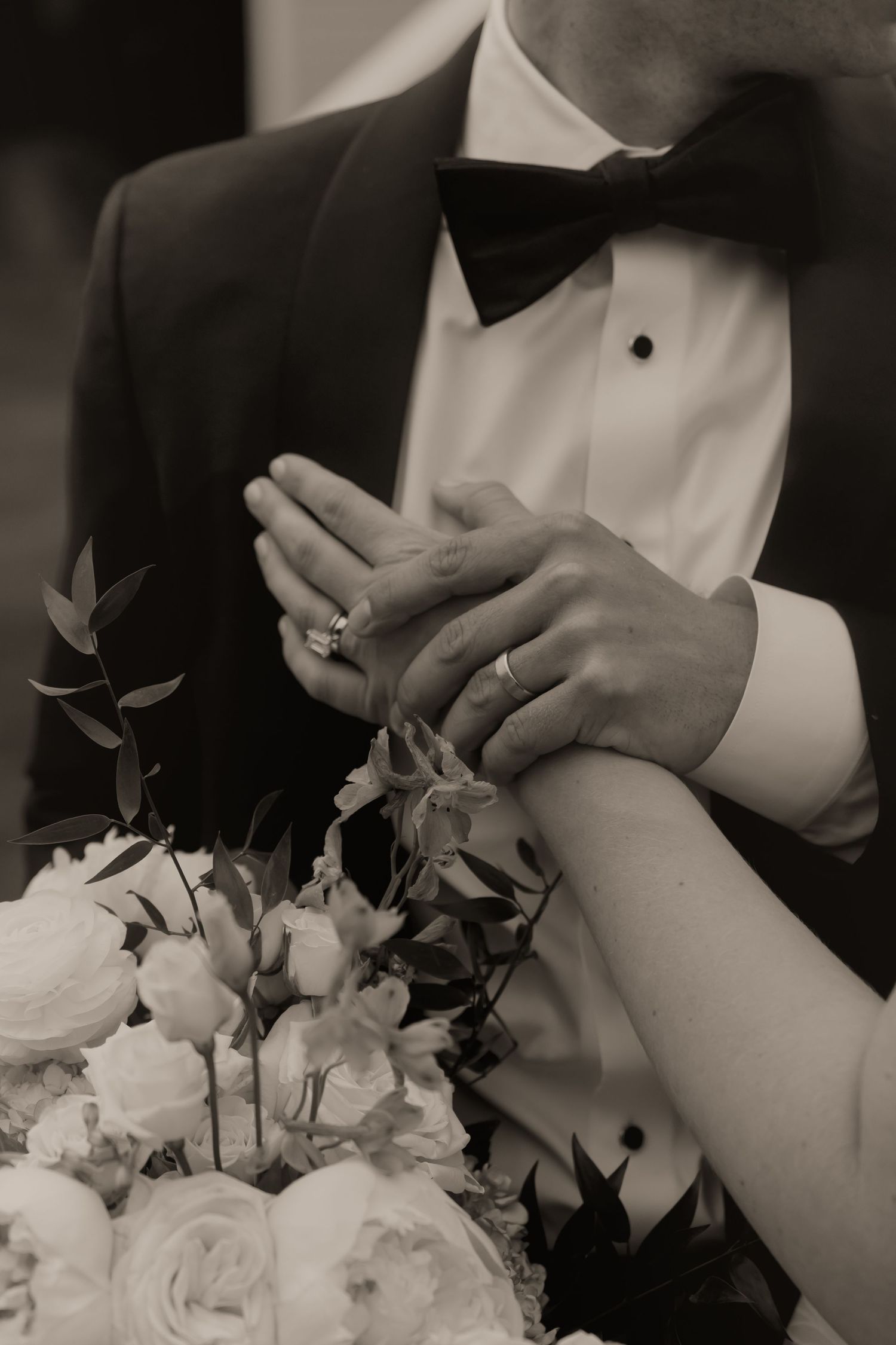Black and white photo of newlyweds holding hands showing wedding rings over bridal bouquet, groom in tuxedo.