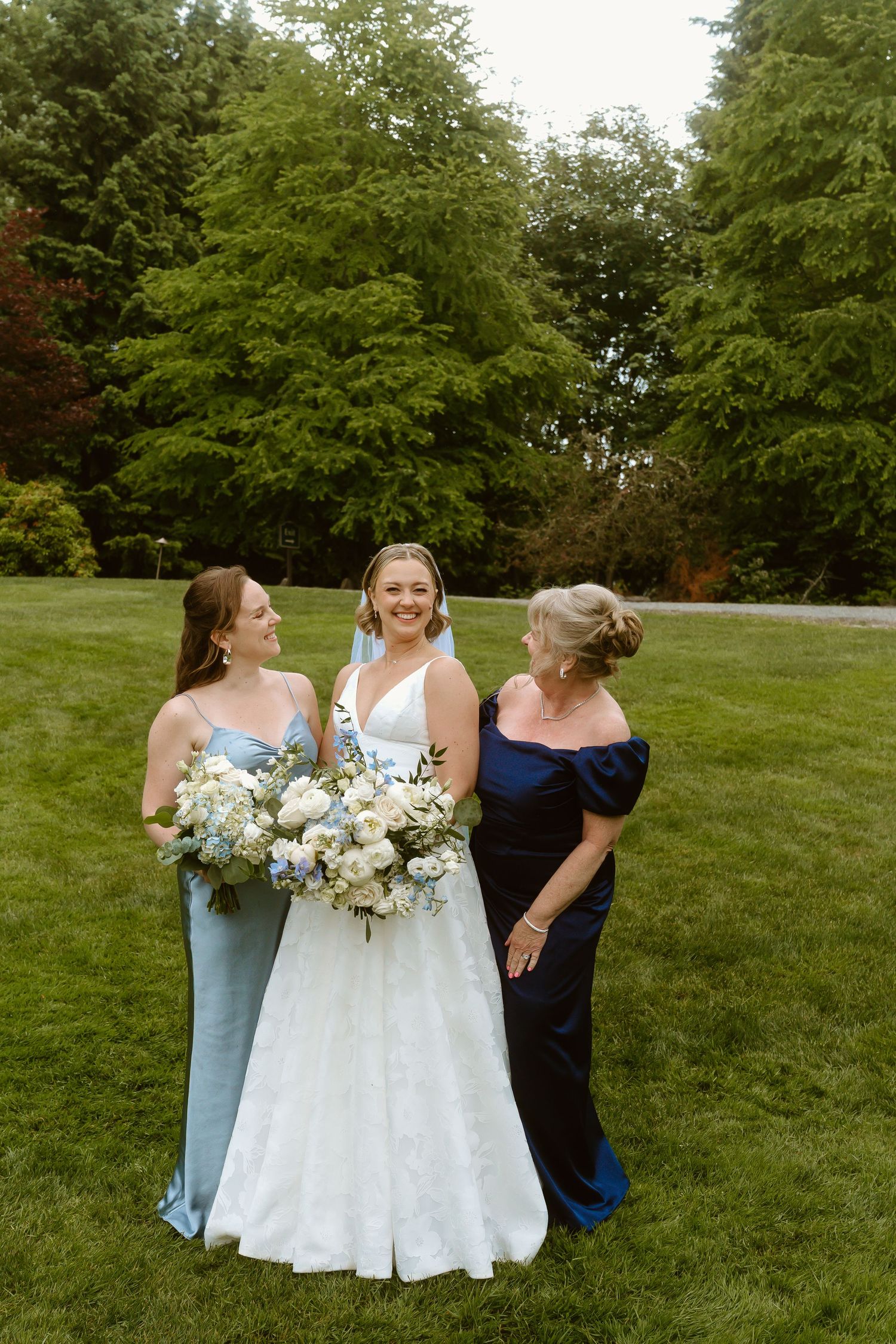 Bride in white gown with veil stands between bridesmaid in blue and mother in navy on green lawn.