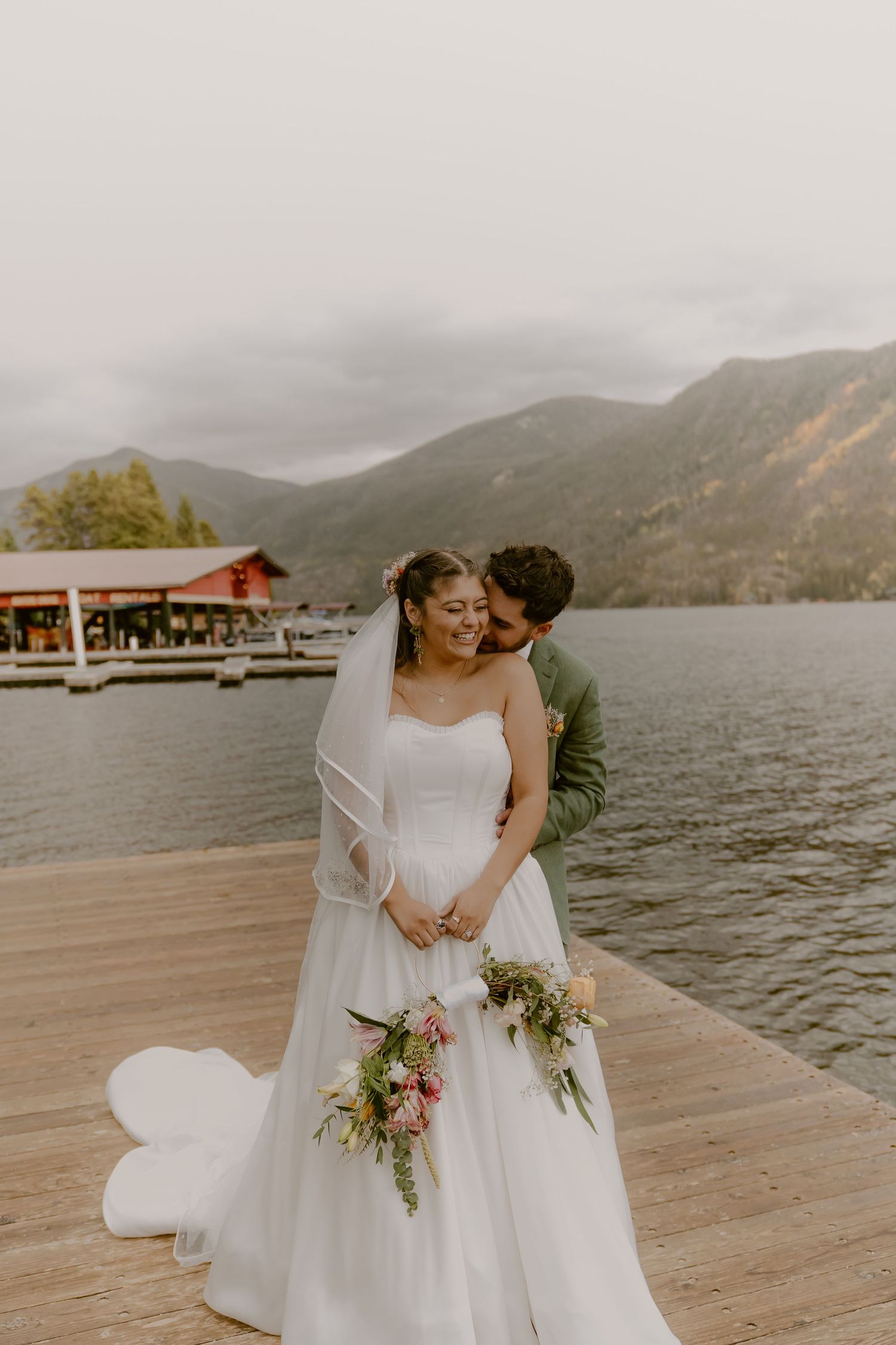 Romantic wedding couple embraces on a lakeside dock with misty mountains in the background.
