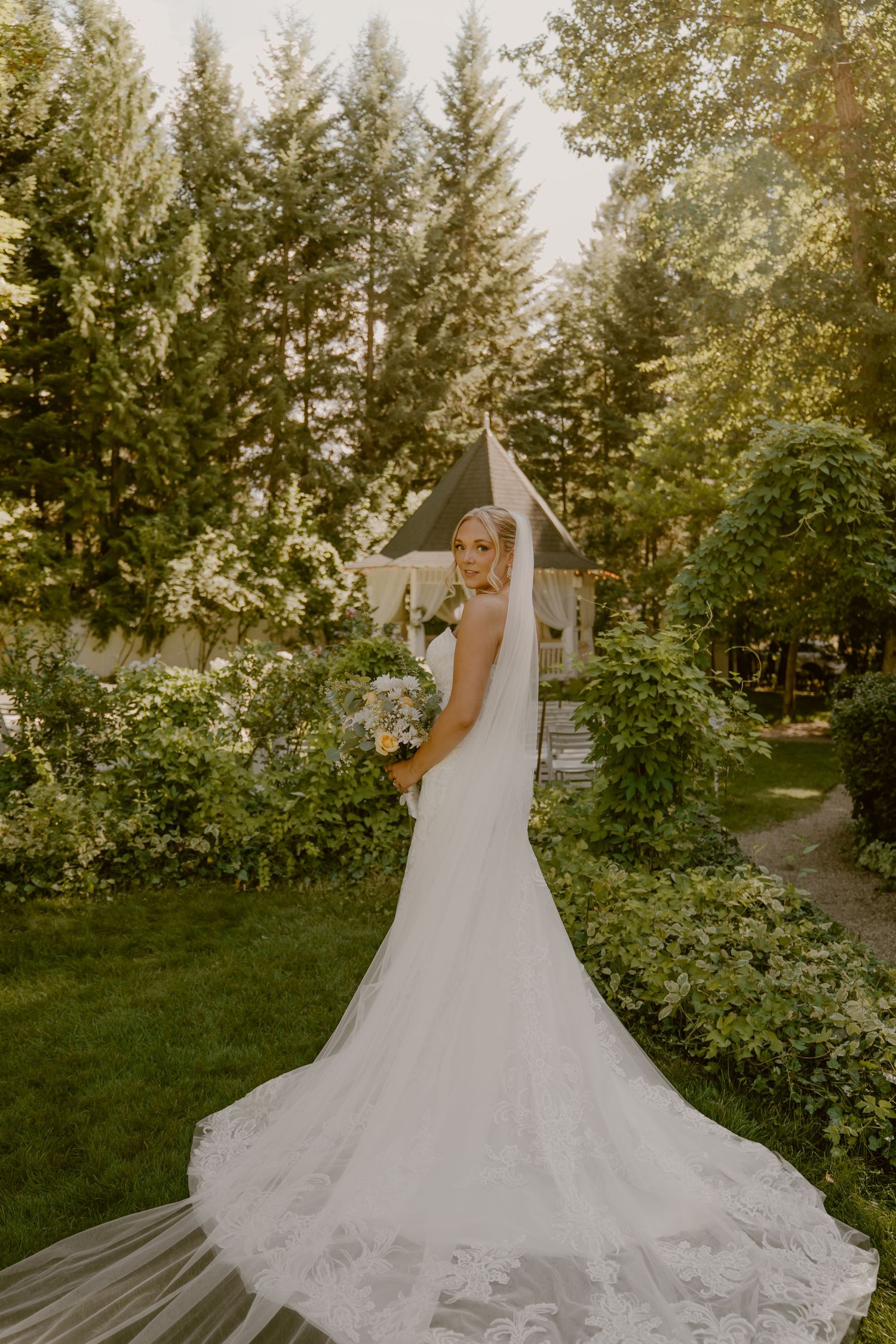 Beautiful wedding dress with long lace train photographed in garden setting with gazebo and evergreen trees.