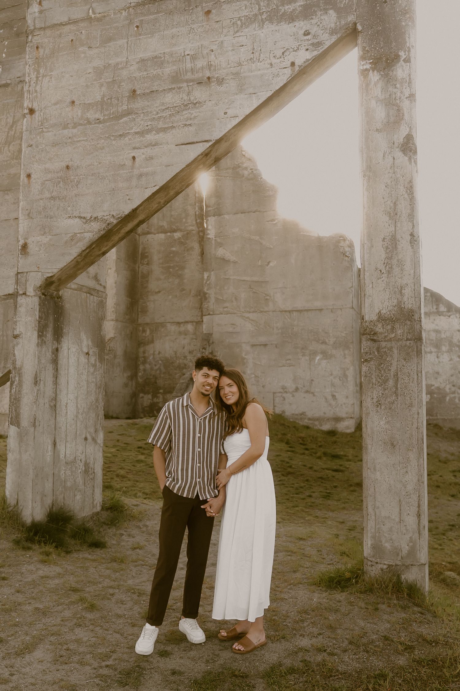 Couple posing romantically near concrete ruins at golden hour, woman in white dress, man in striped shirt.