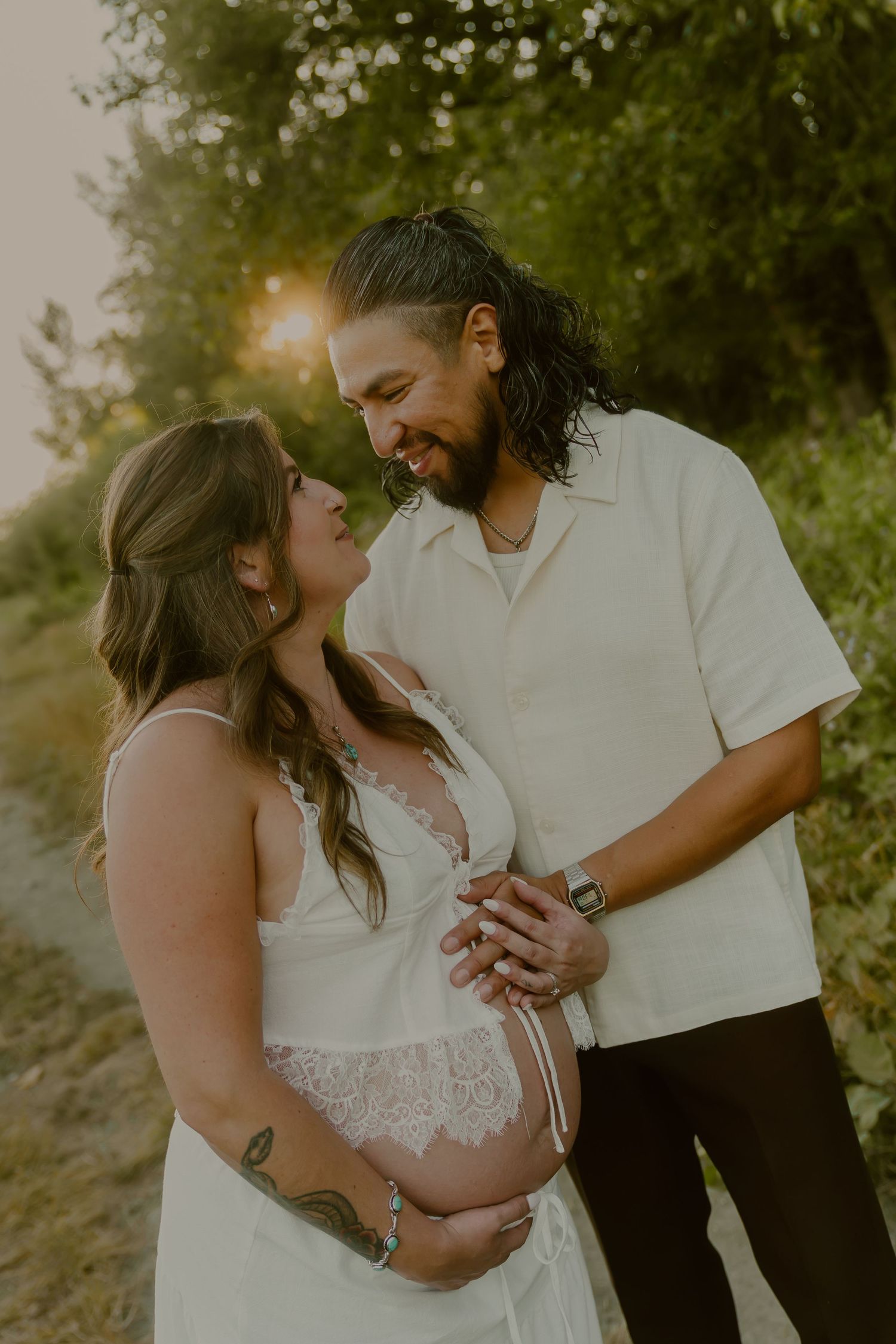 Couple smiling at each other during golden hour maternity photo shoot outdoors, woman in white lace dress cradling baby bump.