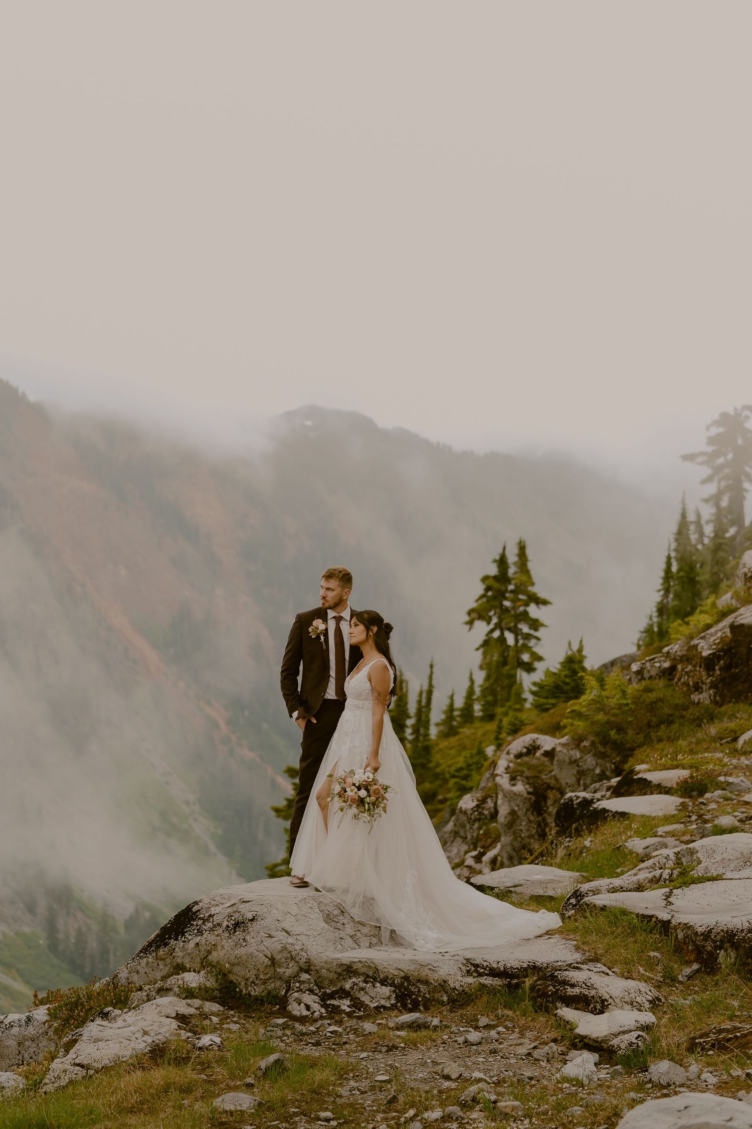 A couple in wedding attire stands on a misty mountain cliff edge surrounded by evergreen trees and dramatic views.