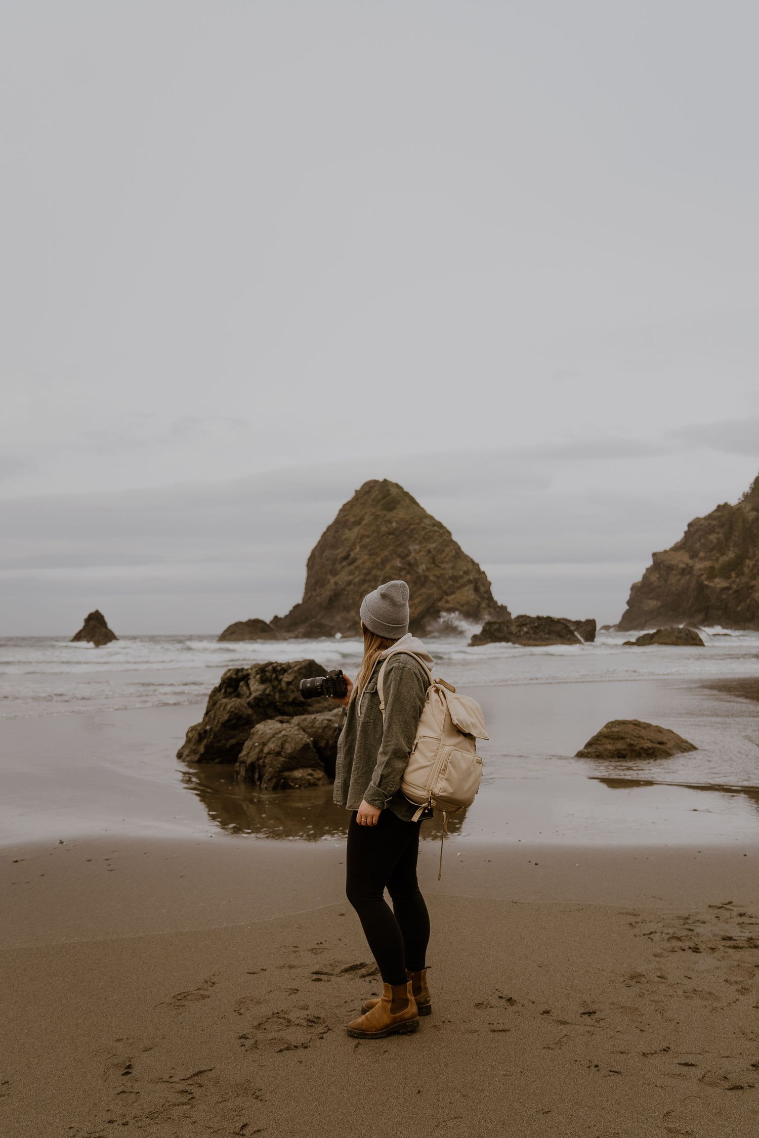 A person in a coat explores a misty beach with large rock formations along the Pacific Northwest coastline.