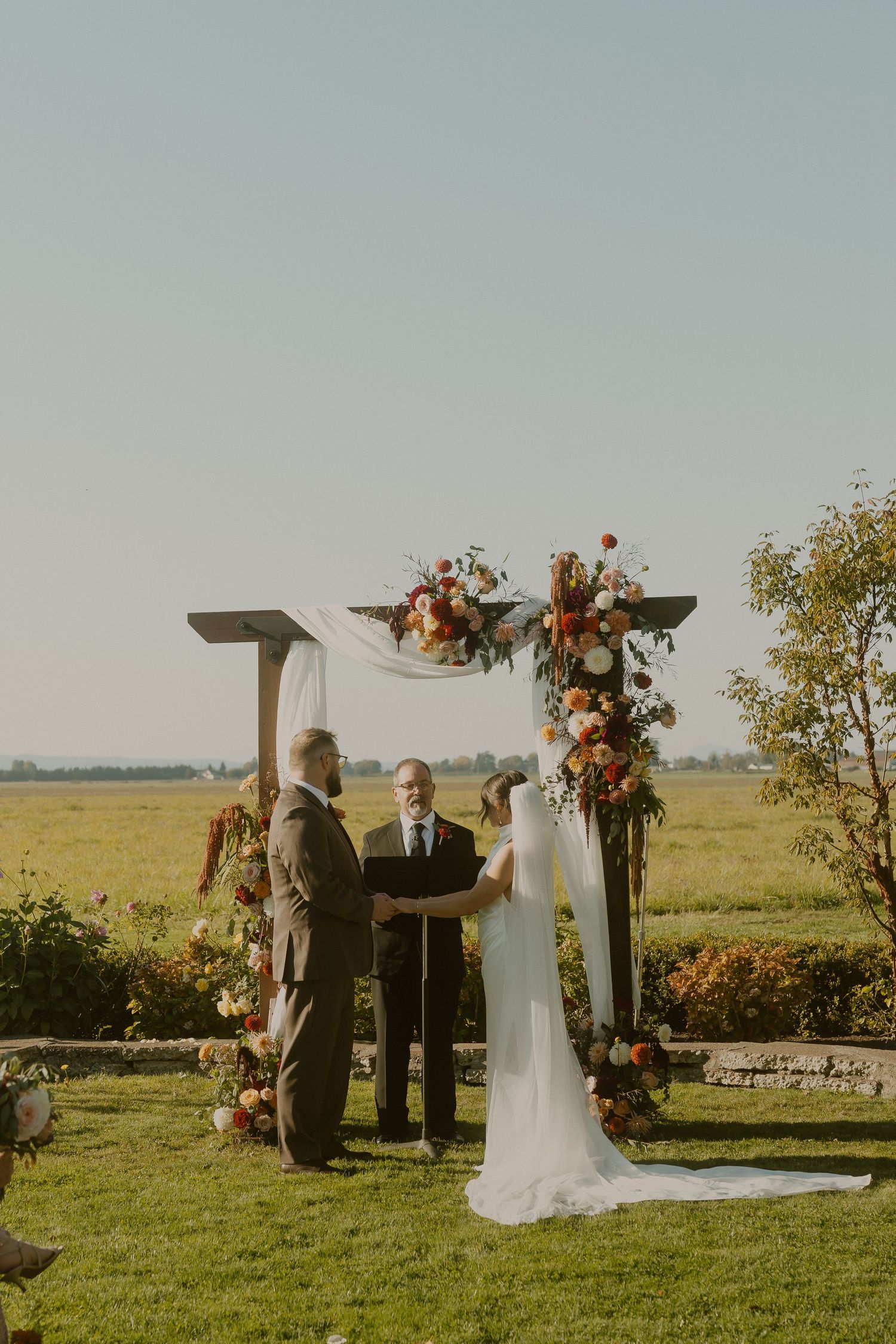Bride in a white gown and groom hold hands before an officiant under a wooden floral arch during an outdoor wedding ceremony in a field.