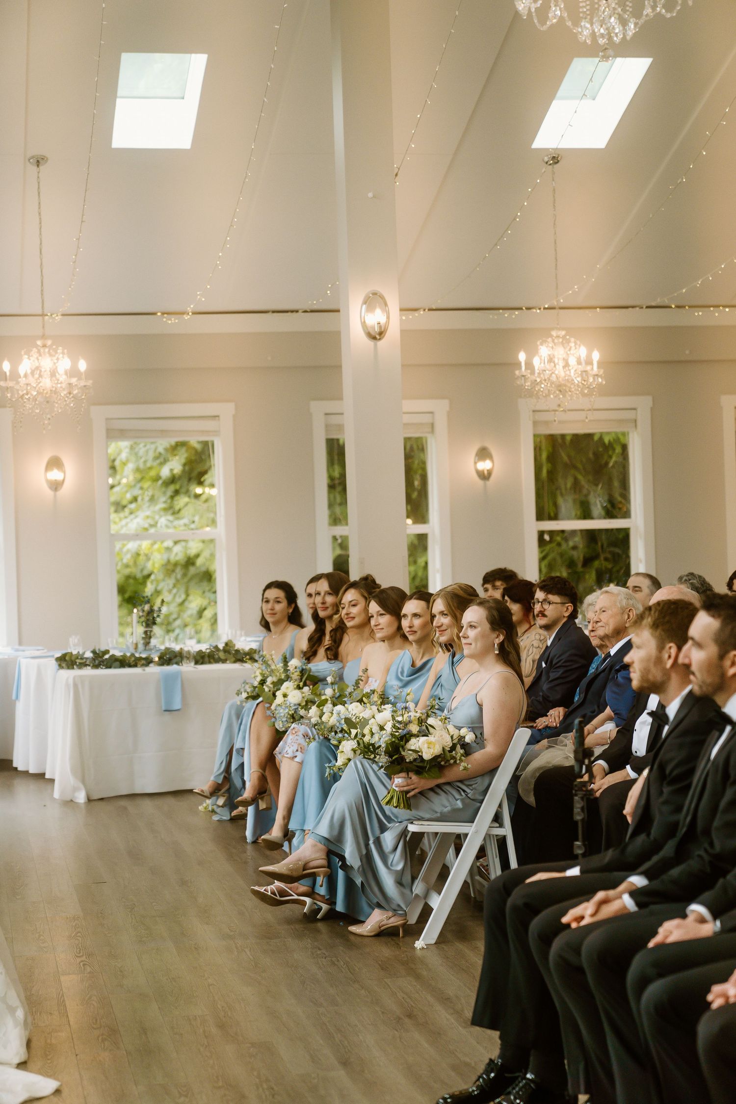 Bridesmaids in blue dresses seated in white chairs during an elegant indoor wedding ceremony with chandeliers.