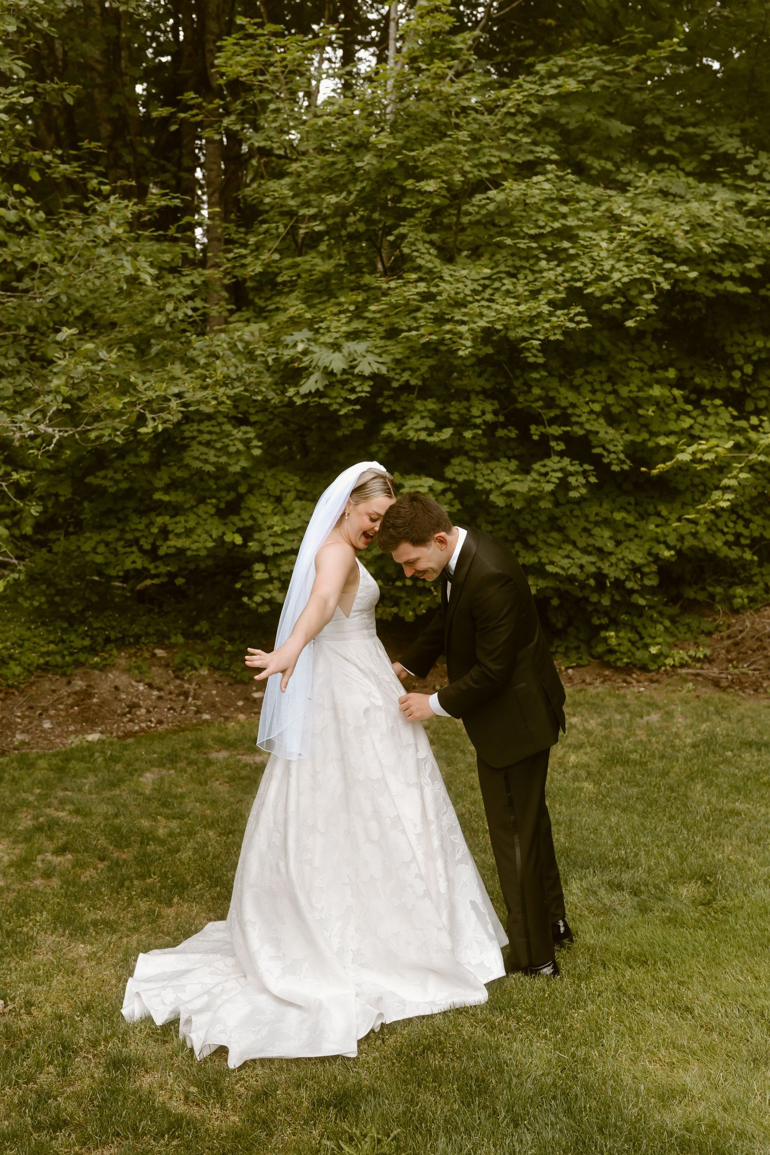 Bride and groom share a tender moment on green lawn, groom bowing toward bride in white gown.