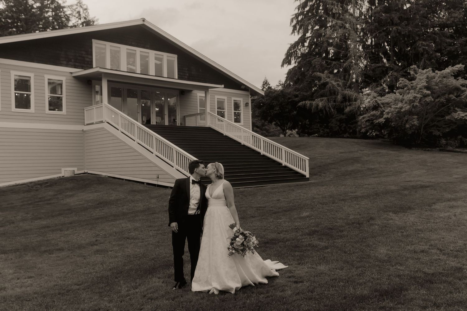 Black and white photo of bride and groom kissing on lawn before elegant white venue with grand staircase.
