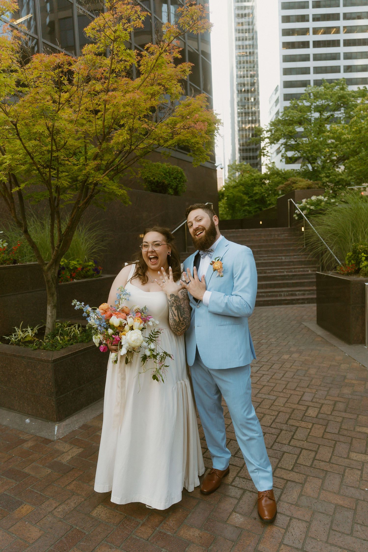 A joyful bride and groom pose outdoors in an urban setting, she in white holding a colorful bouquet, he in a light blue suit.