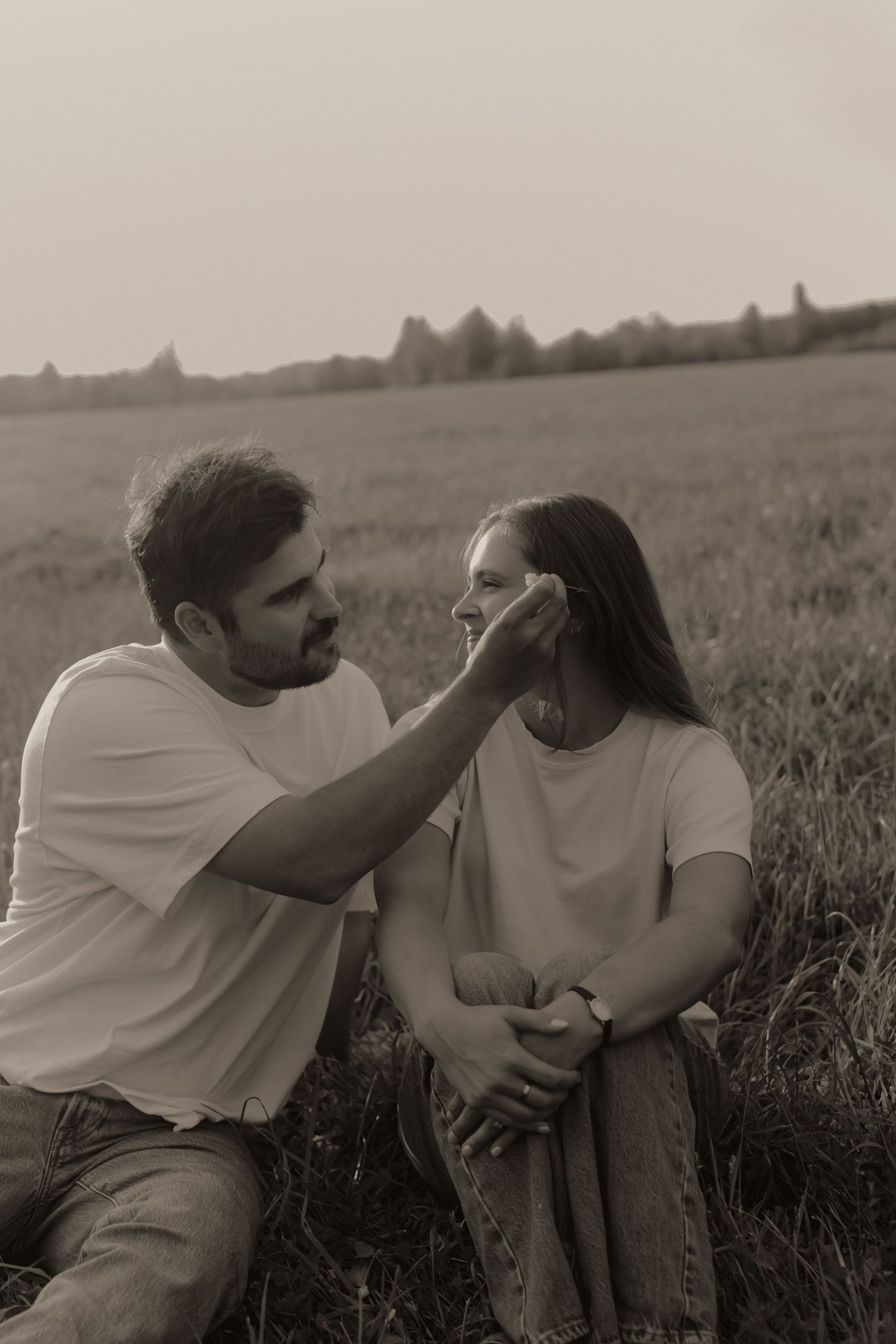 Black and white photo of man gently brushing hair from womans face while sitting in a field.
