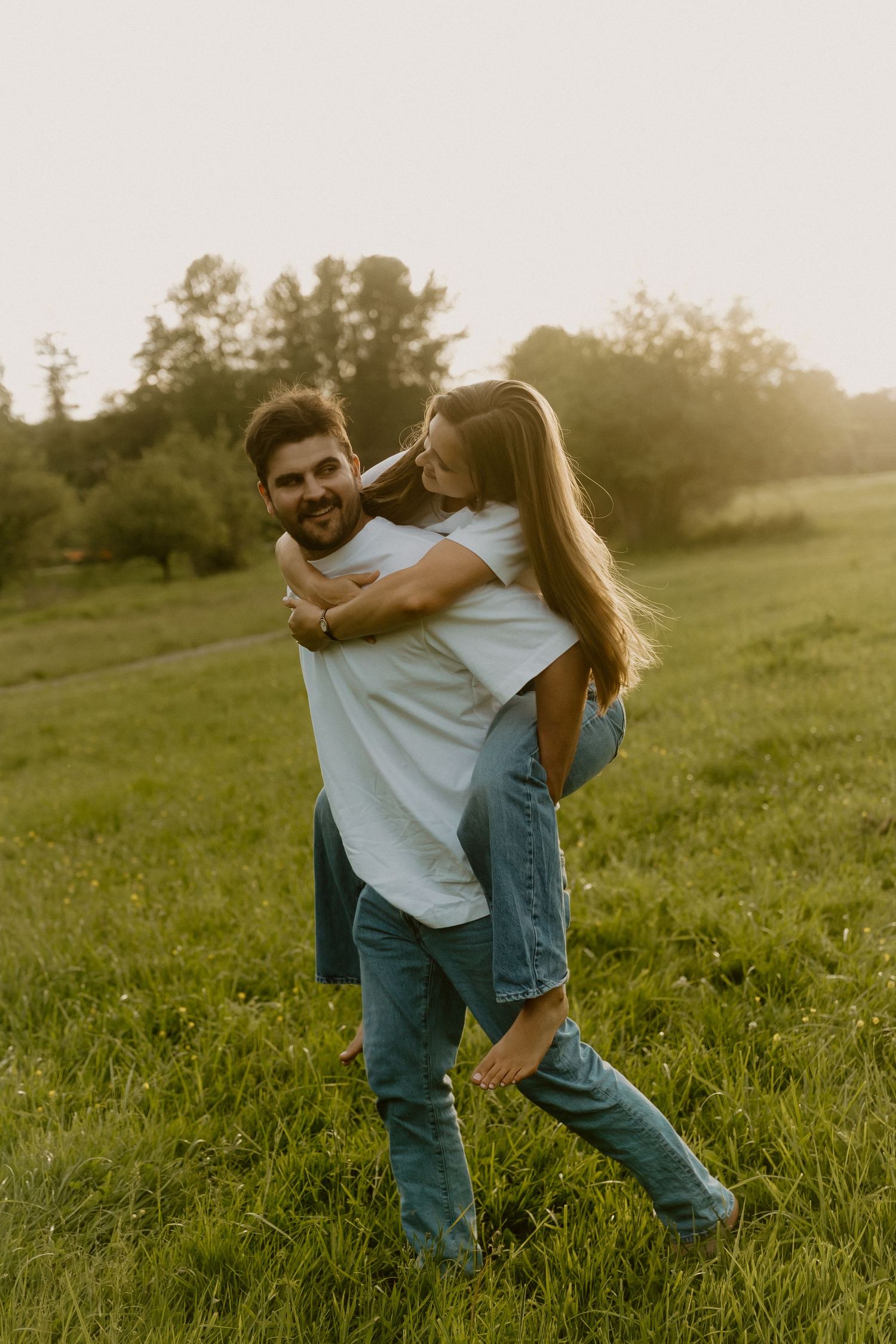 Man giving woman a piggyback ride in a sunny green field, both wearing white shirts and jeans.